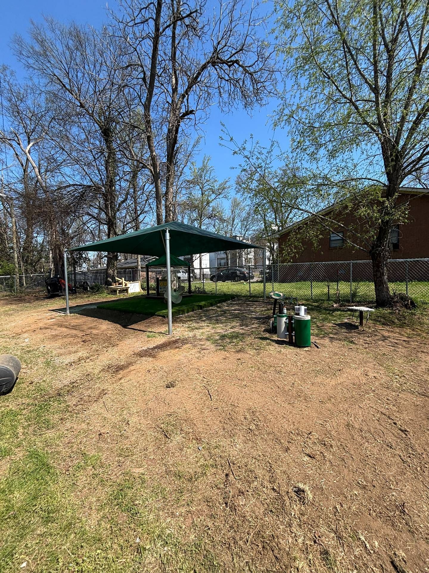 Park shelter with green roof, picnic tables, and trash cans in a grassy area with leafless trees under a blue sky