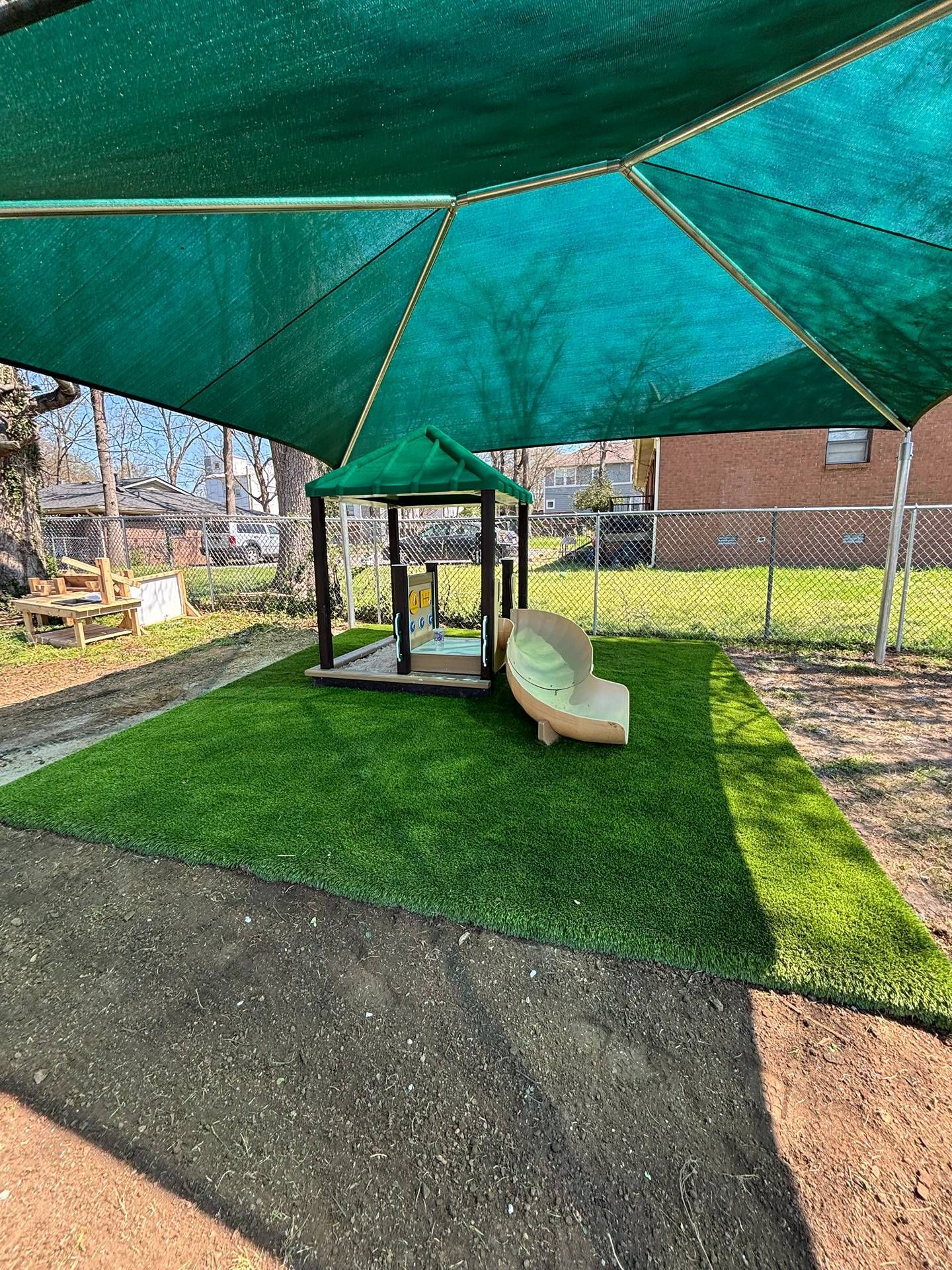 Backyard playground under a large green shade sail, with a slide and swing set on turf.