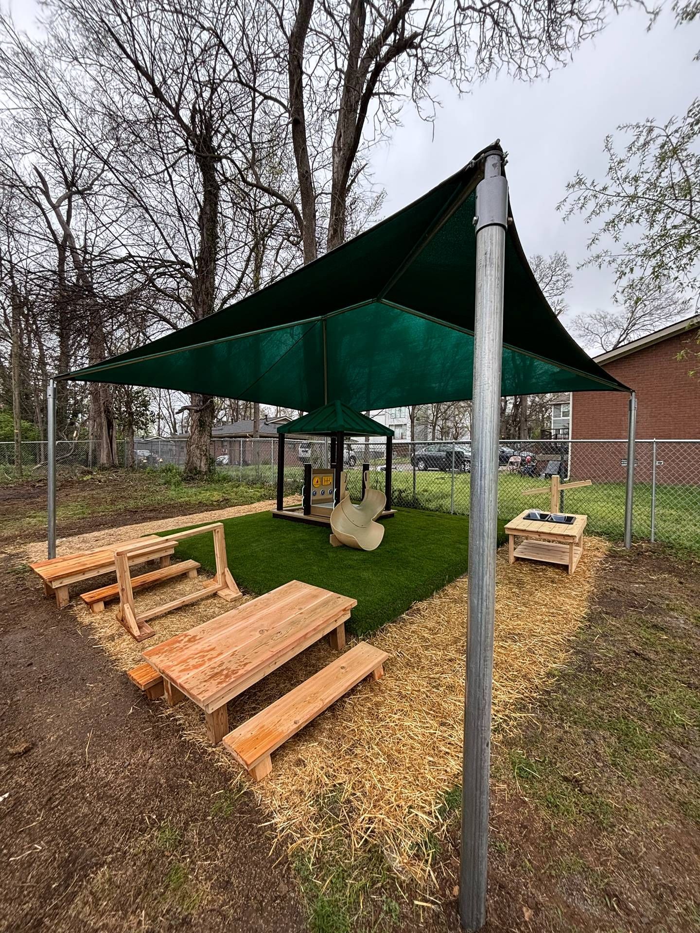 Outdoor goat pen with green shade sail, wooden benches, and two goats on straw-covered ground.