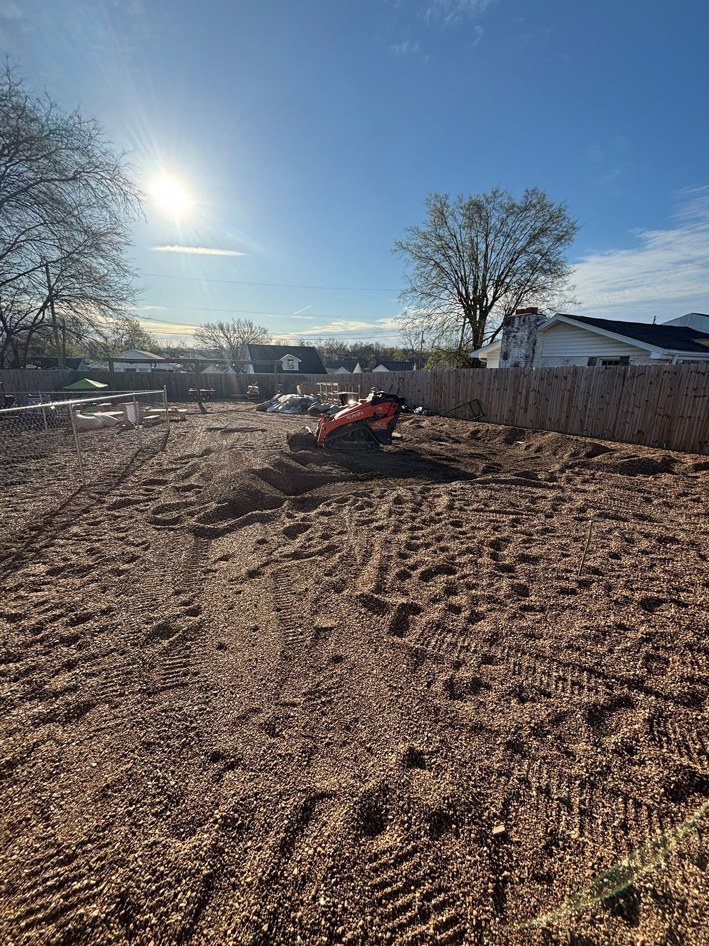 Sunlit rural yard with wood chips, a red wheelbarrow, leafless trees, and a white fence under a blue sky