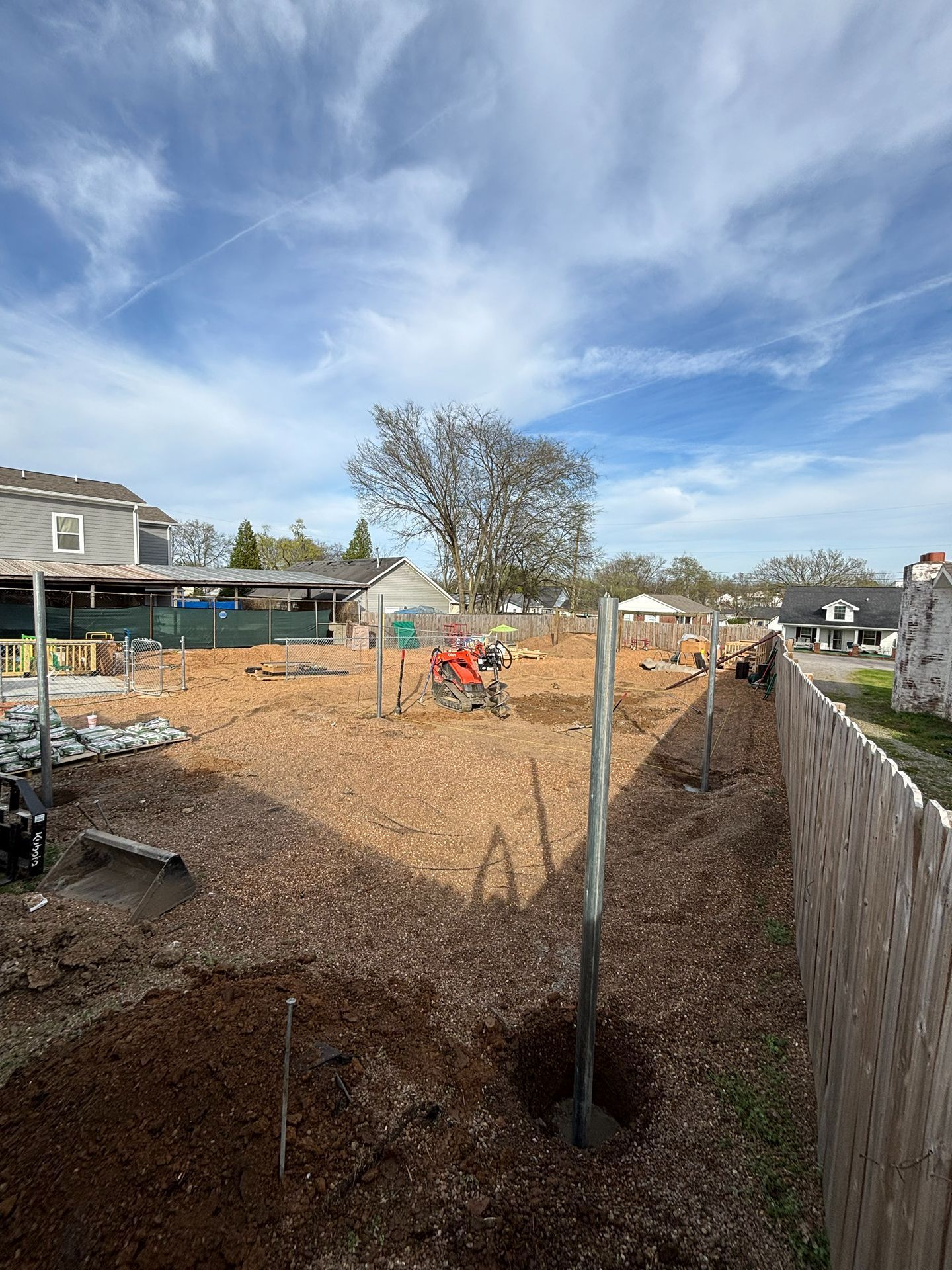 Backyard under construction with fresh excavation, orange equipment, and a concrete retaining wall under a blue sky