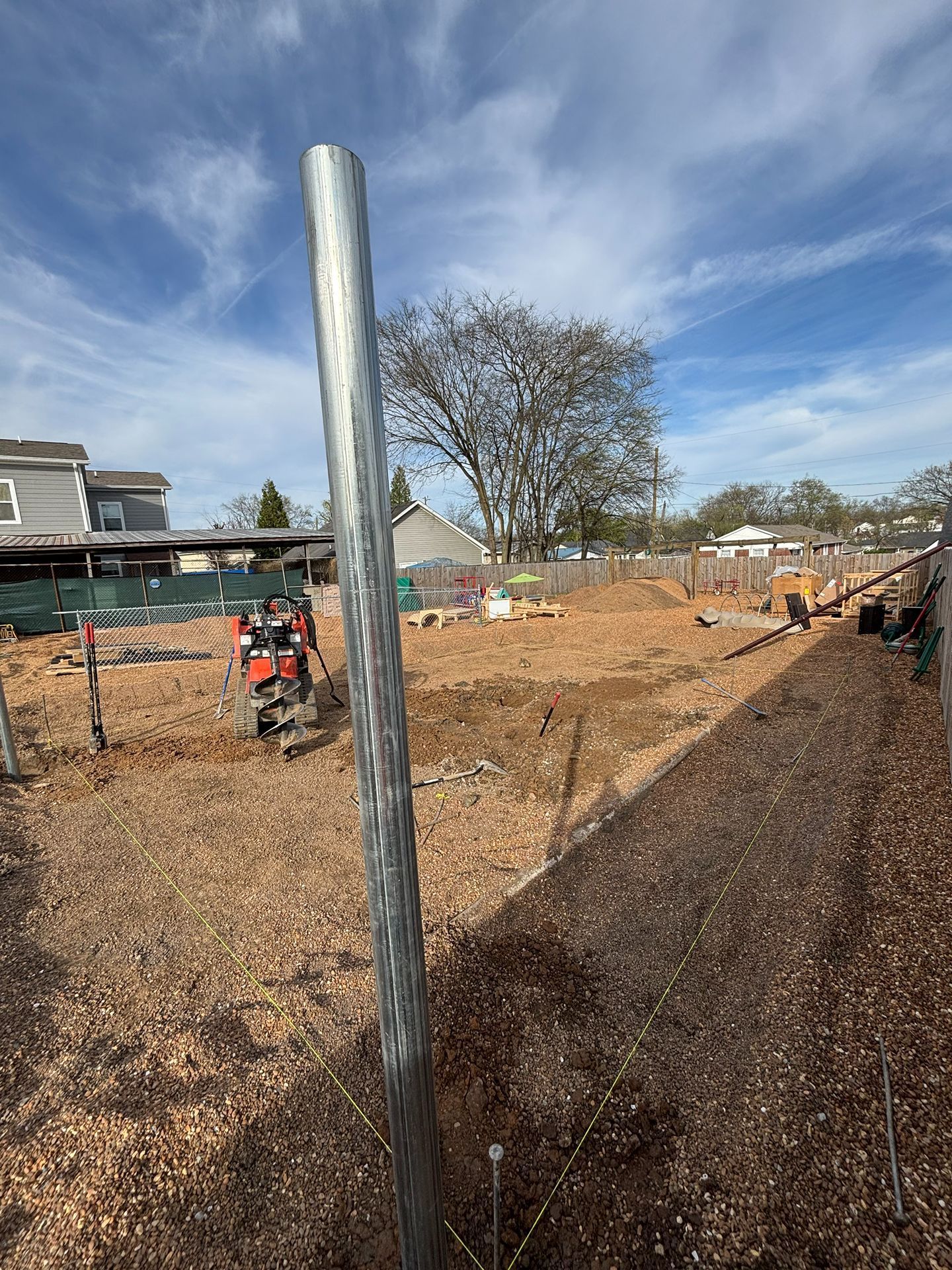A muddy construction site with a tall metal pole, scattered equipment, and a leafless tree under a blue sky.