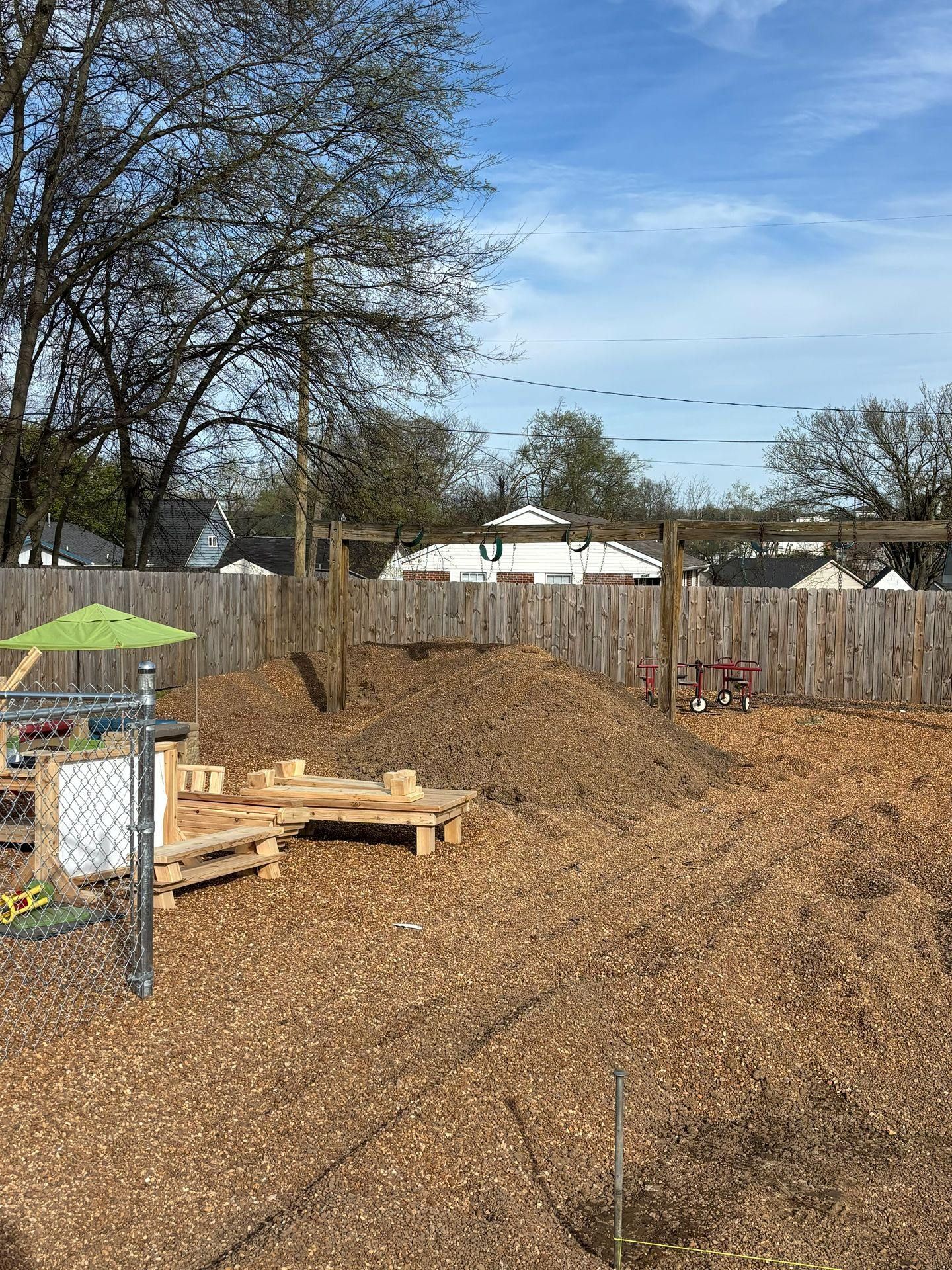Woodchip playground with logs, fence, and bare trees under a blue sky