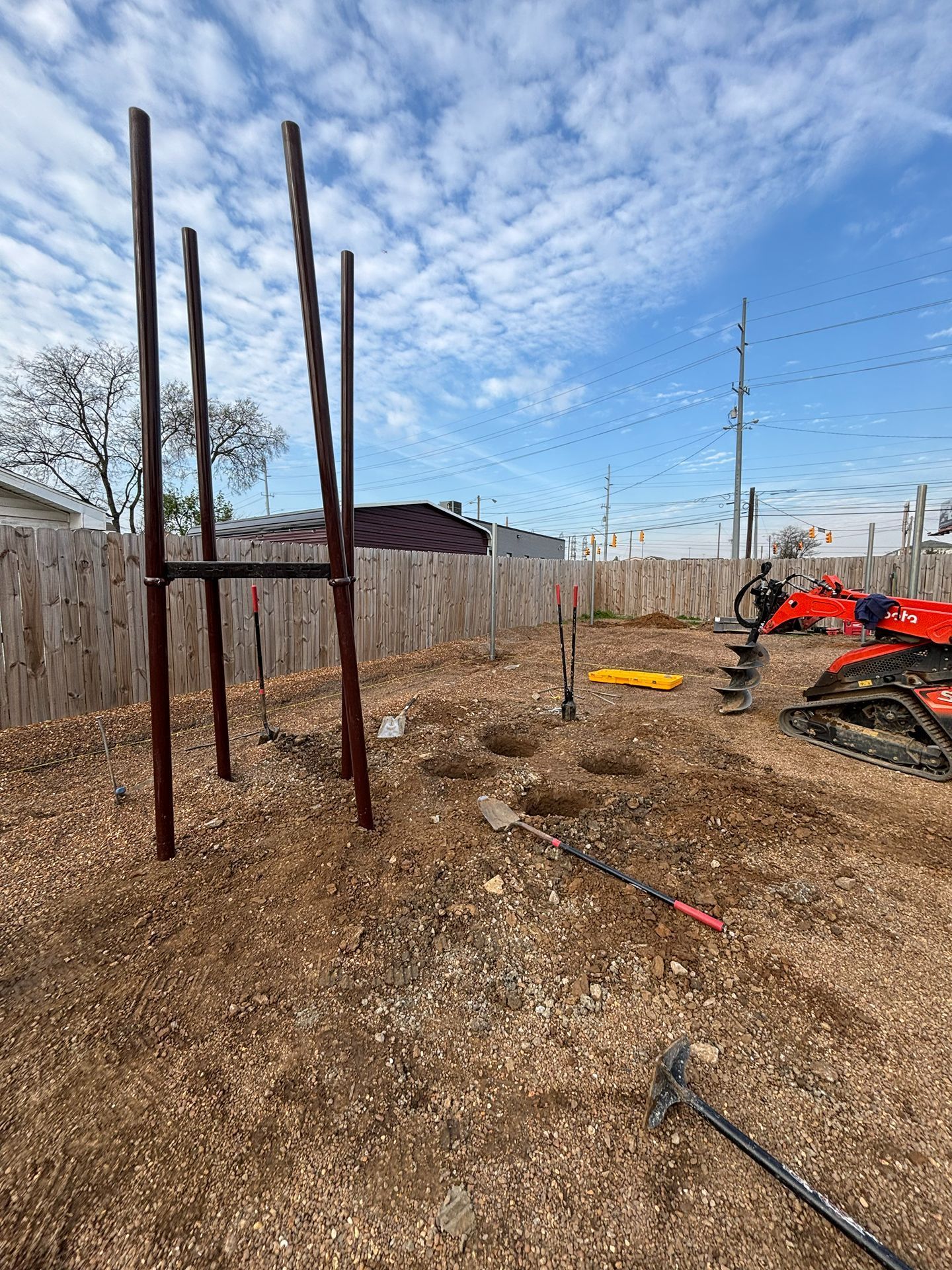 Outdoor yard with tall metal posts, dirt ground, a wooden fence, and a red ATV under a cloudy sky