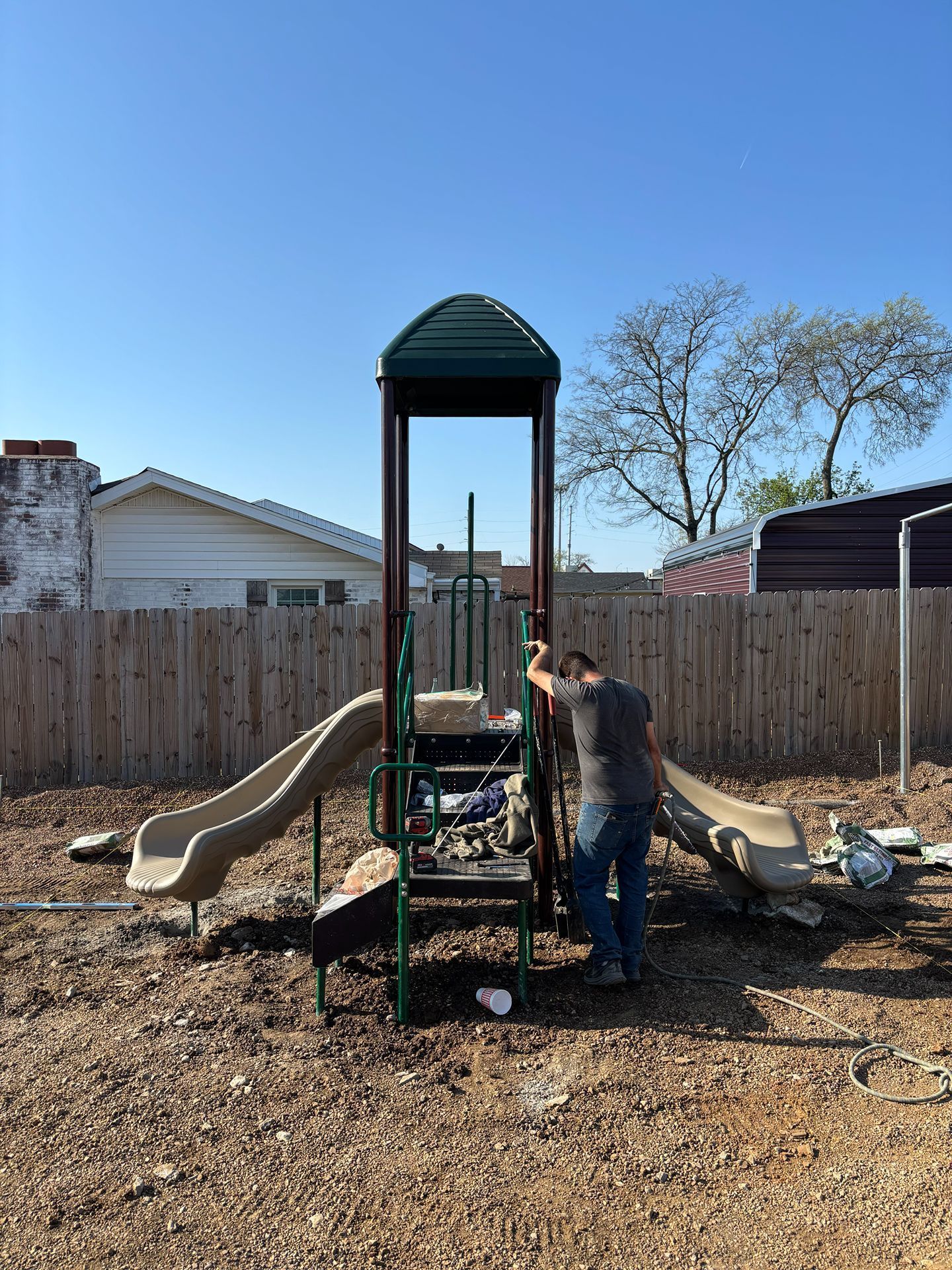 Person working on a playground structure in a backyard under a clear blue sky.