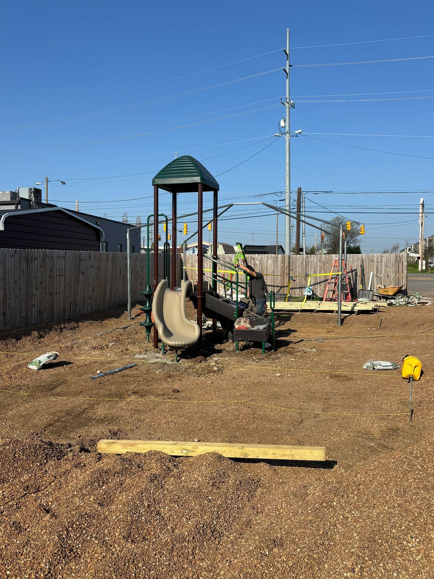 Children’s playground with a slide in a dirt yard, fenced in, under a clear blue sky.