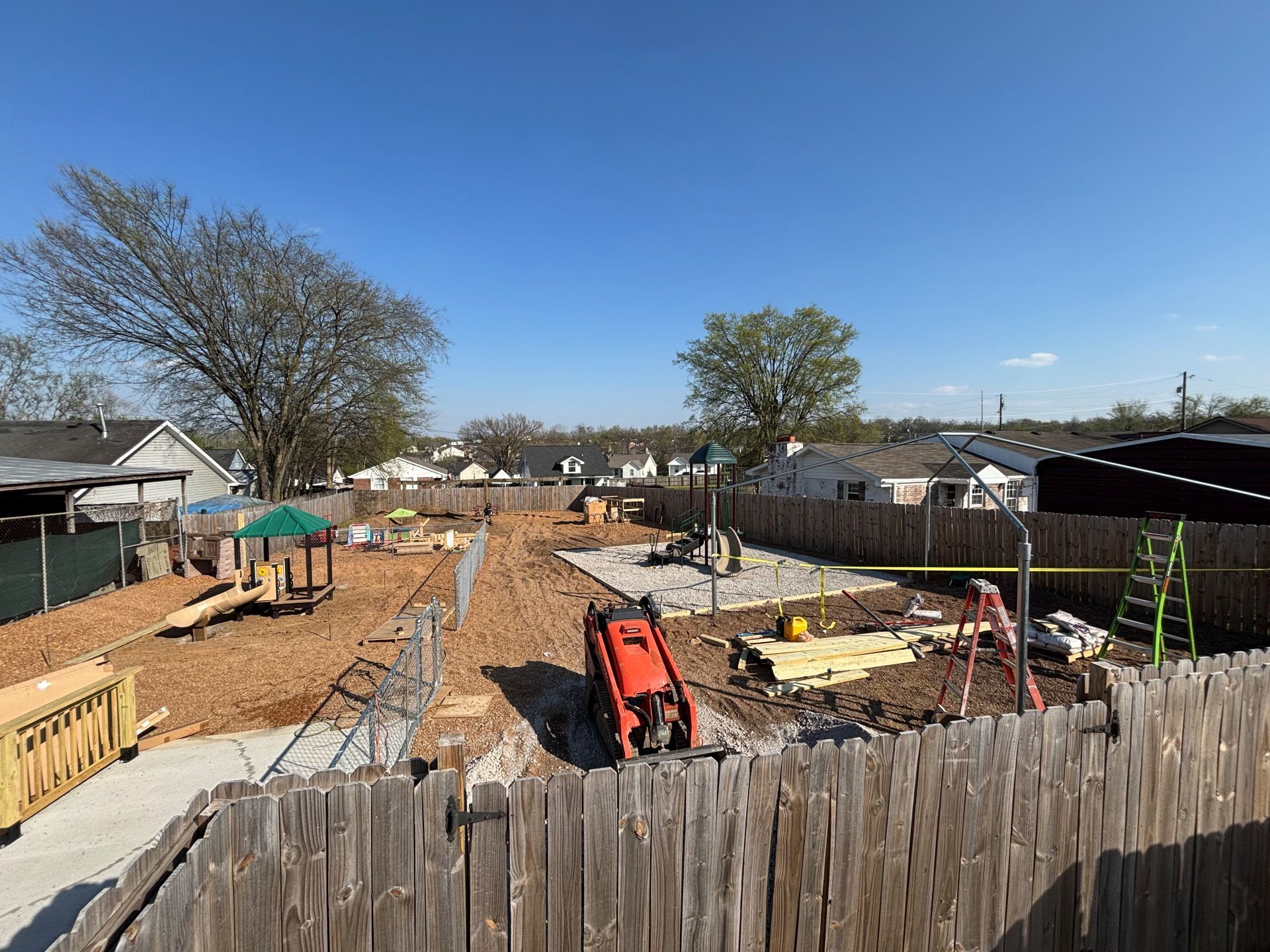 Construction site with wooden foundations, dirt trenches, and equipment in a fenced residential area under a blue sky