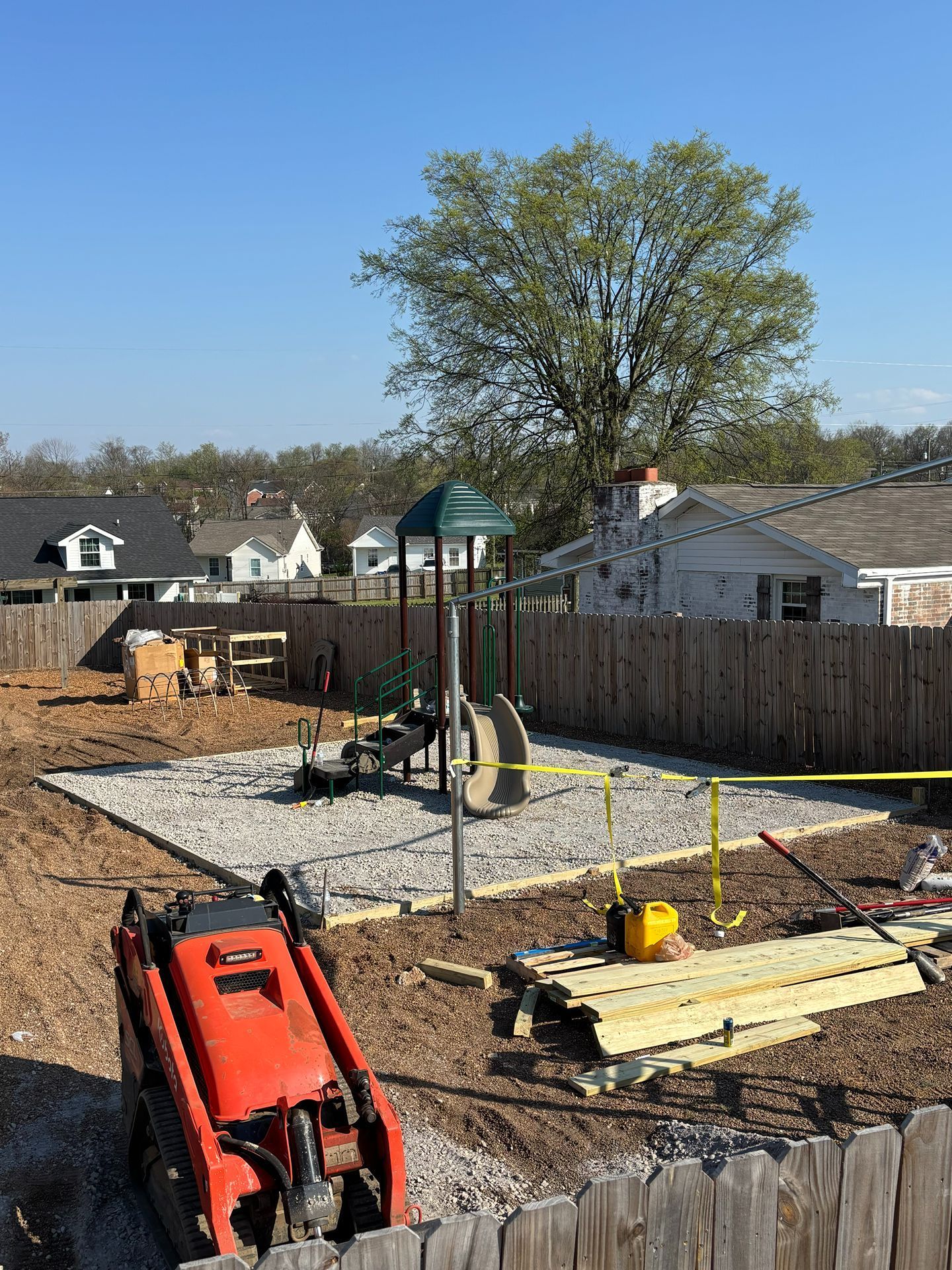 Backyard playground under construction with swings, mulch, fence, and a red excavator in the foreground