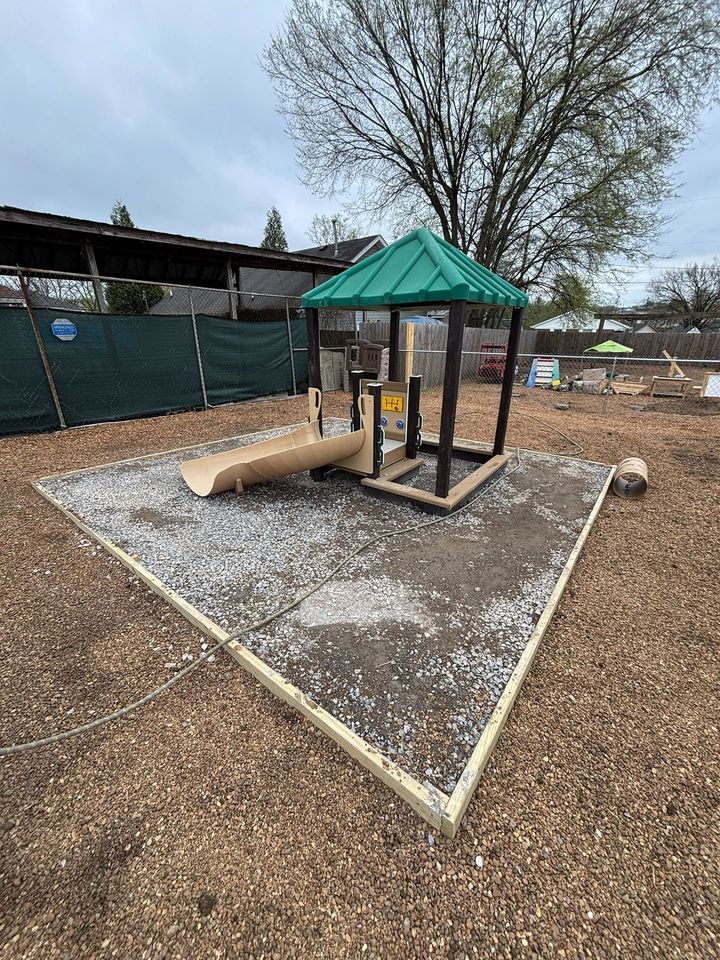 Small playground structure with green roof and slide on a triangular gravel area under construction.