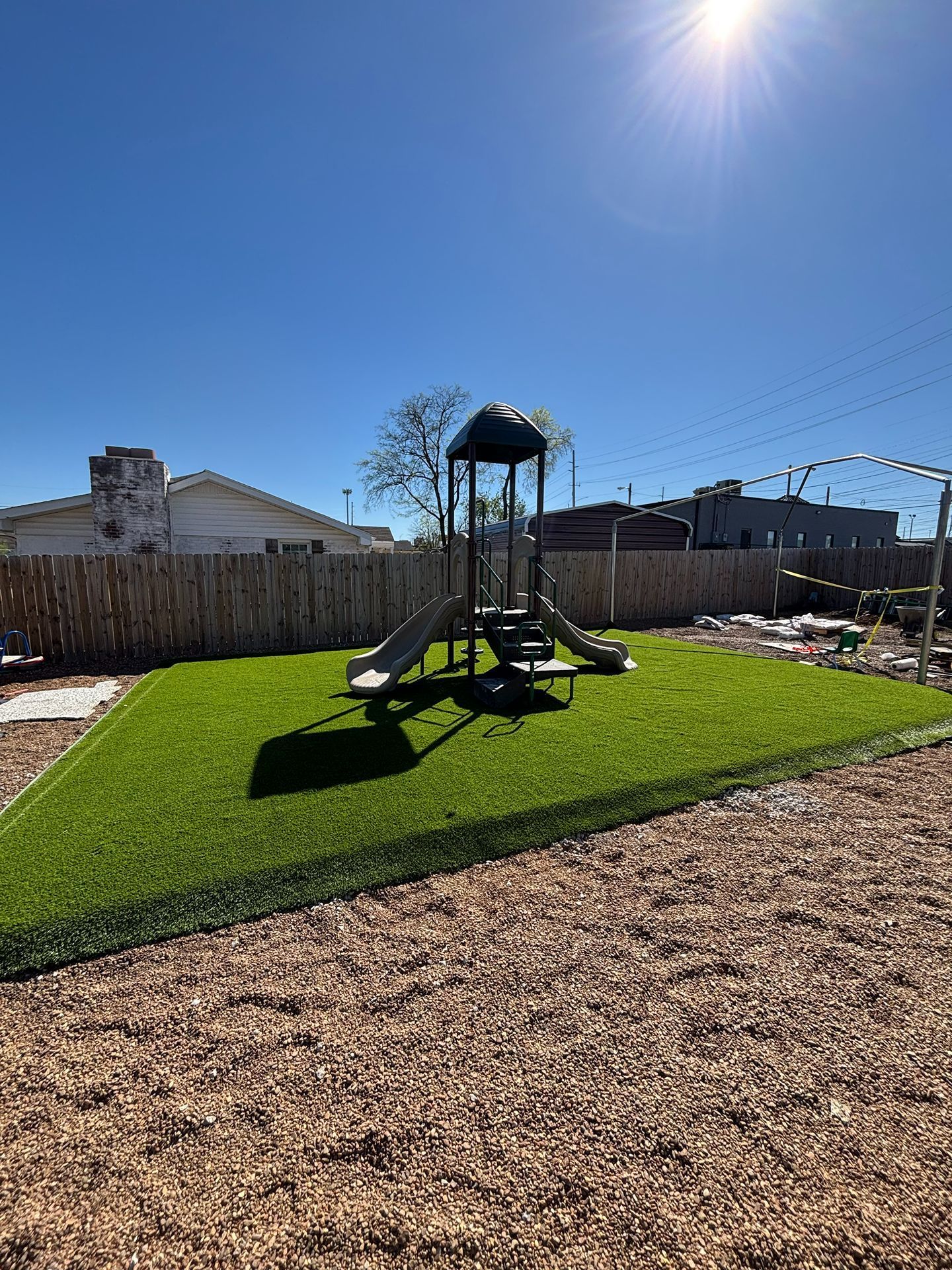 Sunny playground with green turf, slide, and climbing structure on a mulch-covered yard