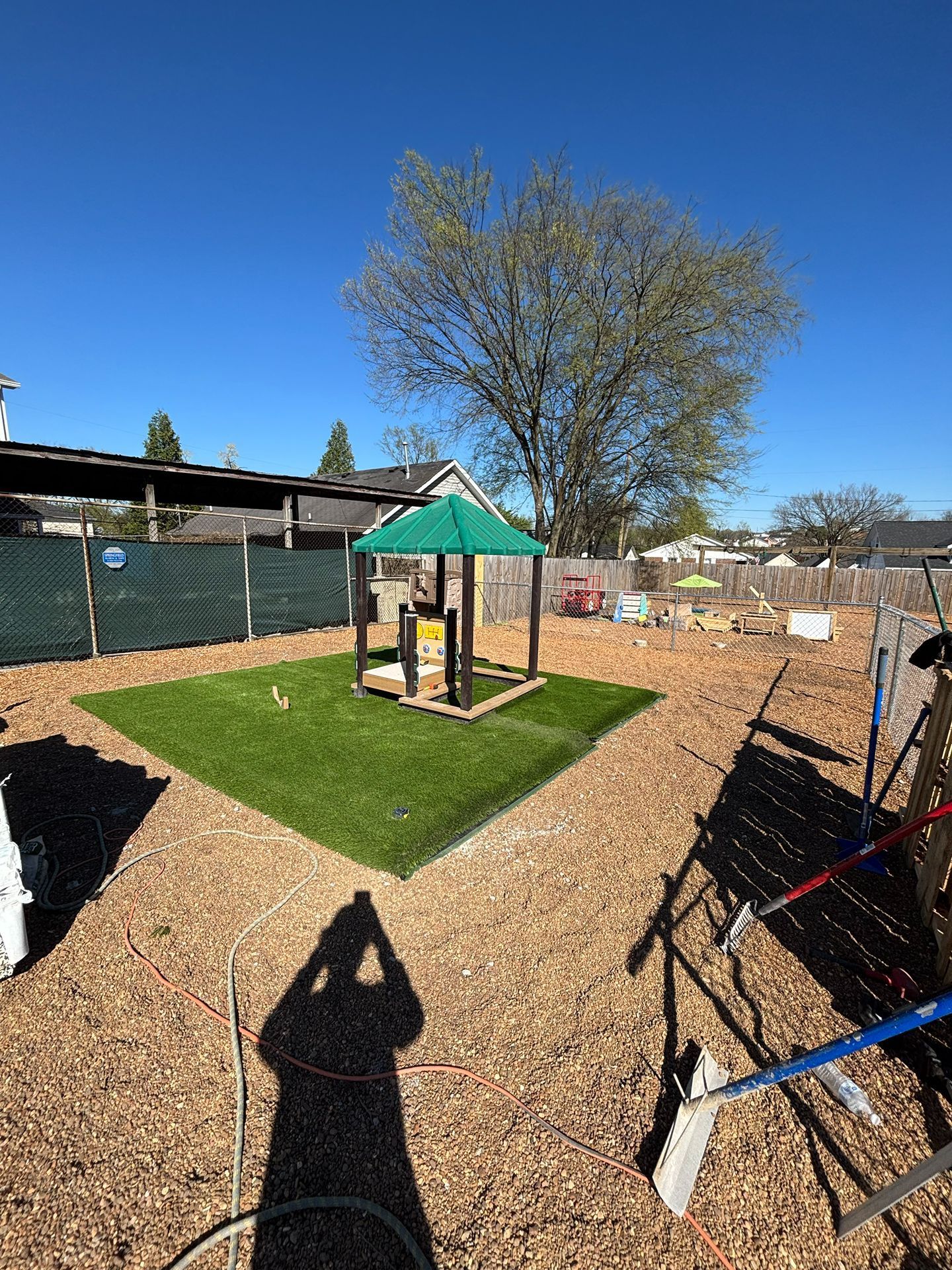 Backyard play area with a small wooden playhouse on a green turf square under a tree, fenced by gravel paths.
