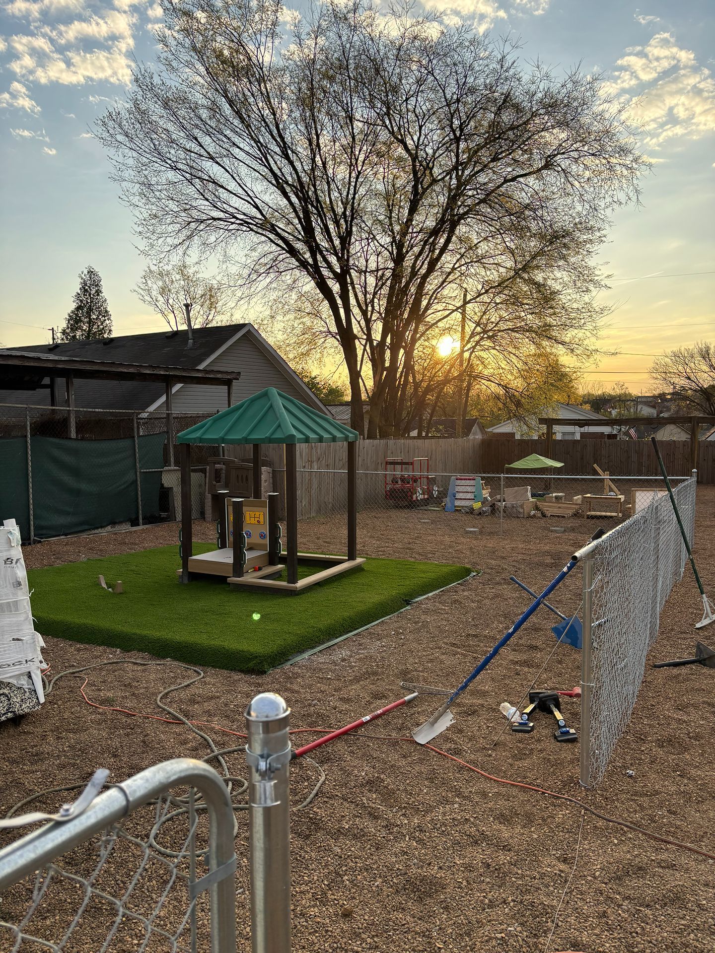 Backyard playground with a small gazebo, fenced area, and a large tree at sunset