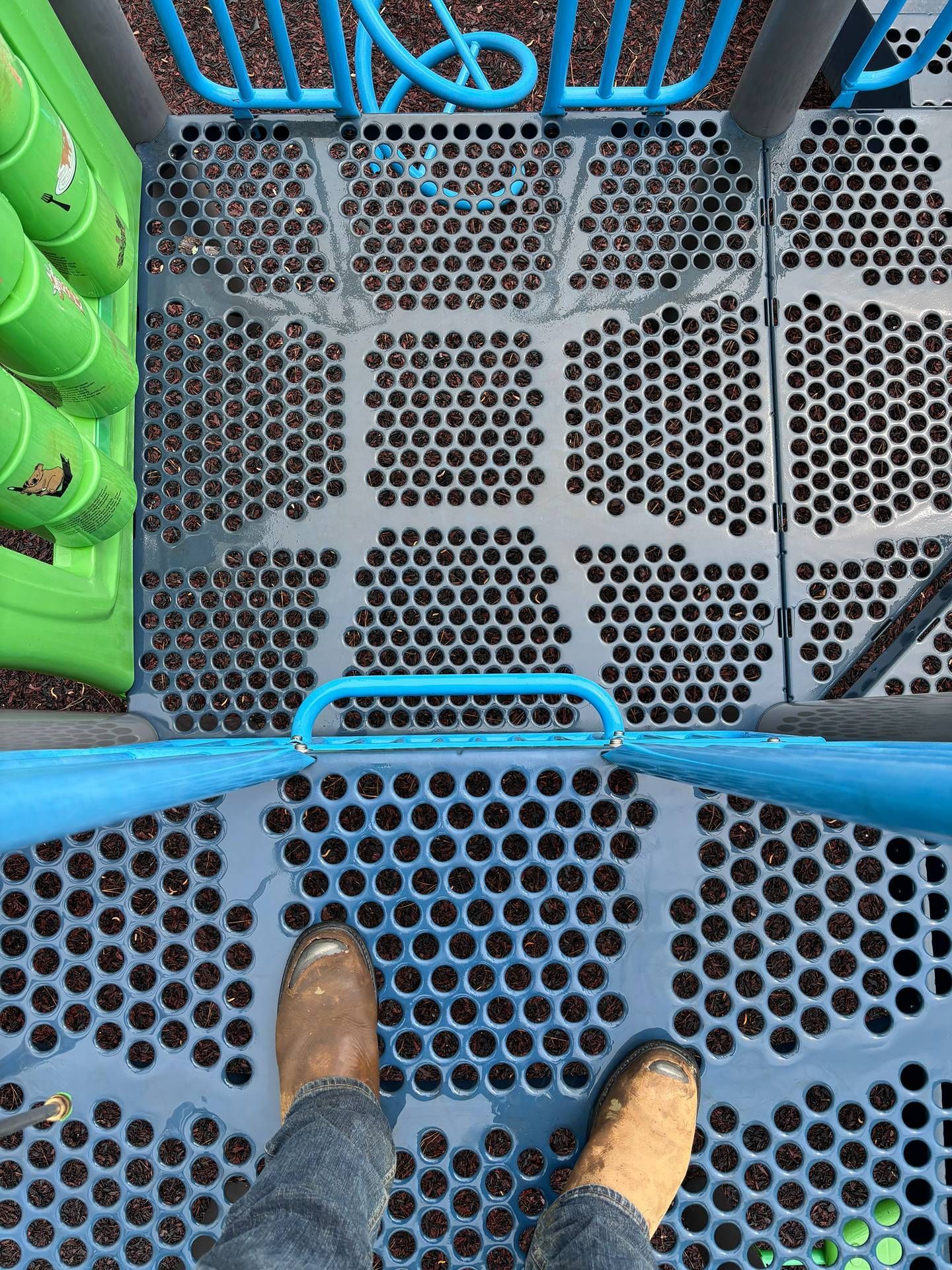Looking down at shoes on a blue perforated metal platform with green and blue playground equipment nearby