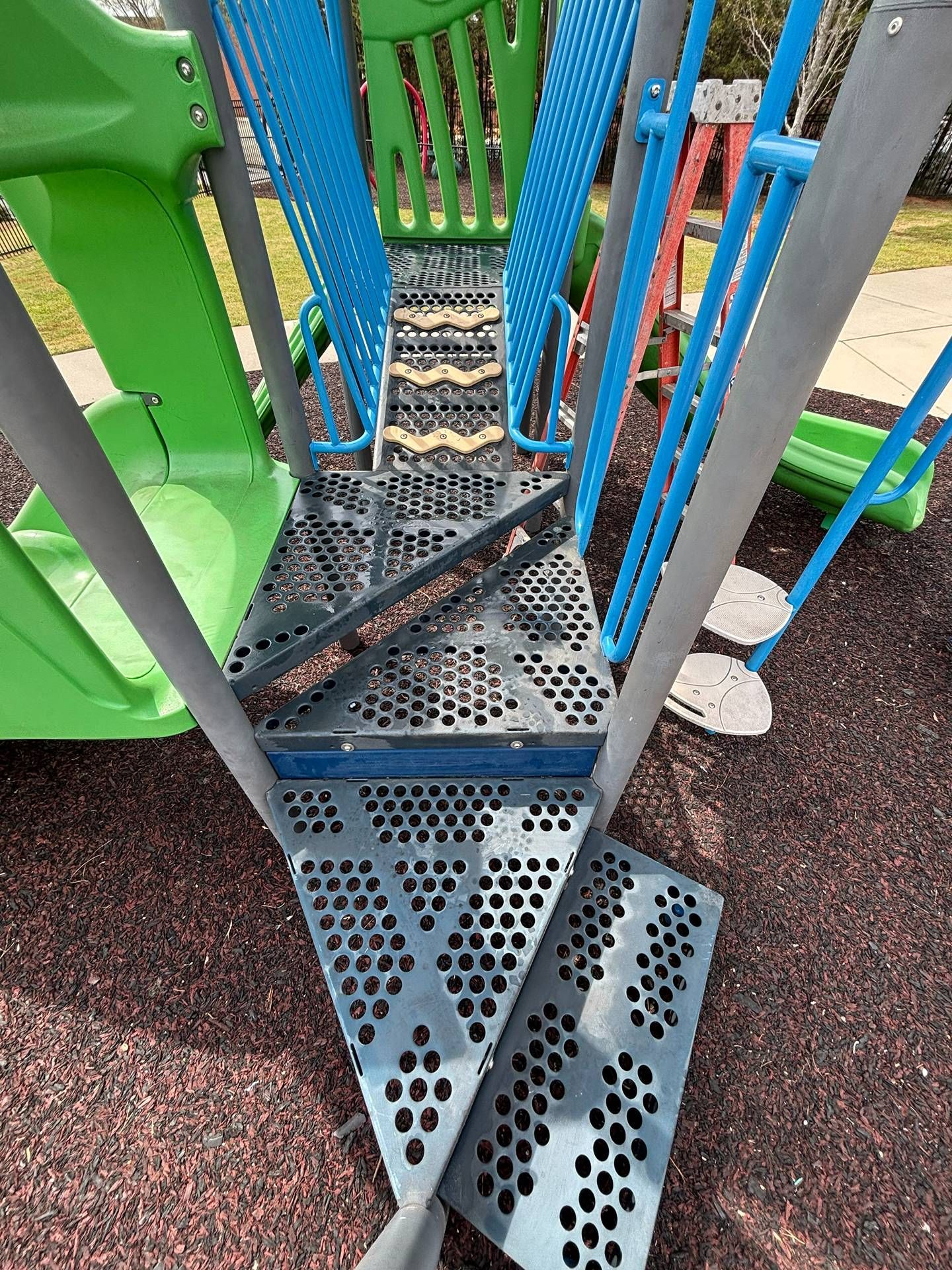 Metal playground staircase with blue and green rails and perforated gray steps.