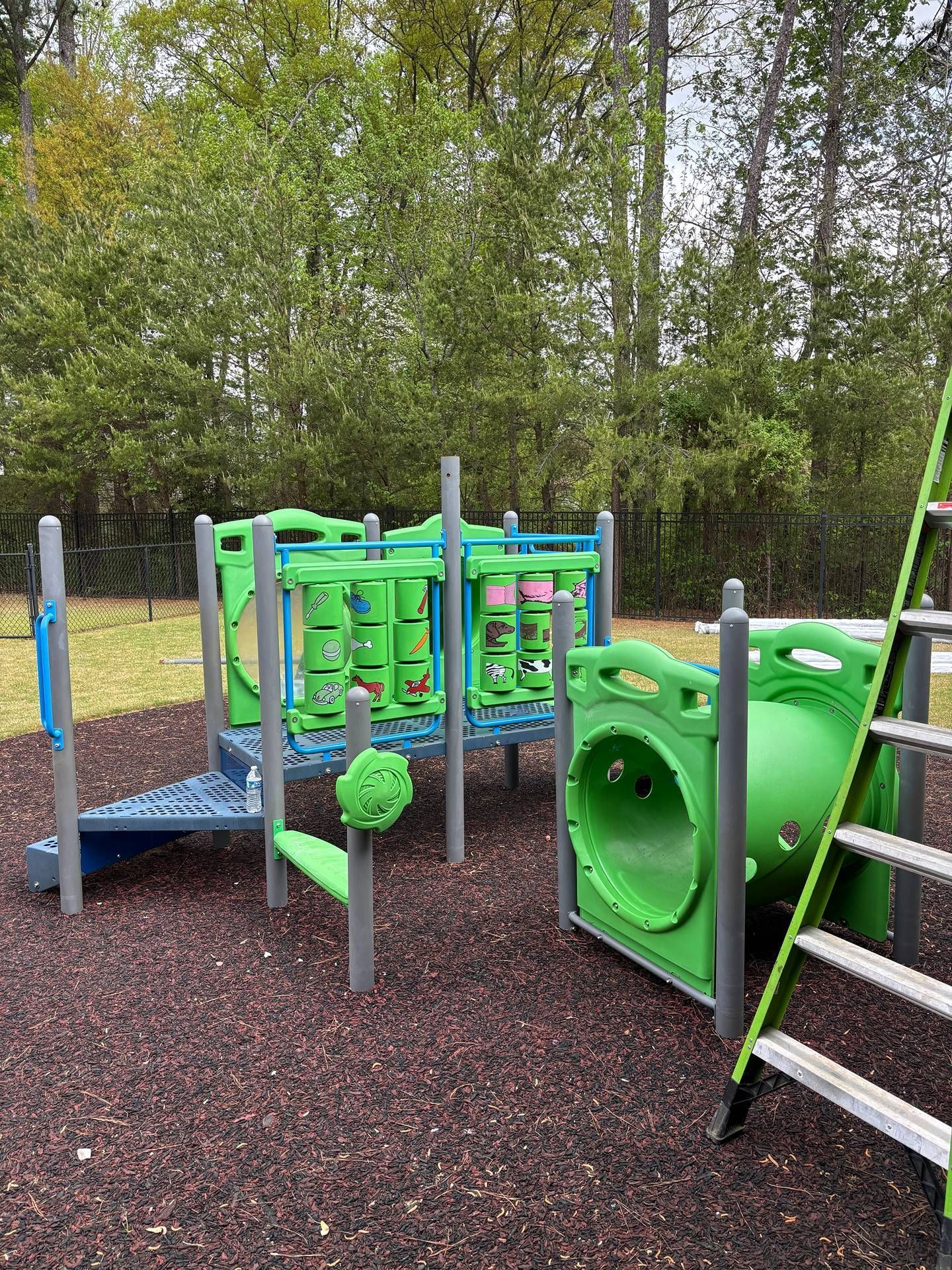 Green playground climbing structure and slide on wood chips beside trees