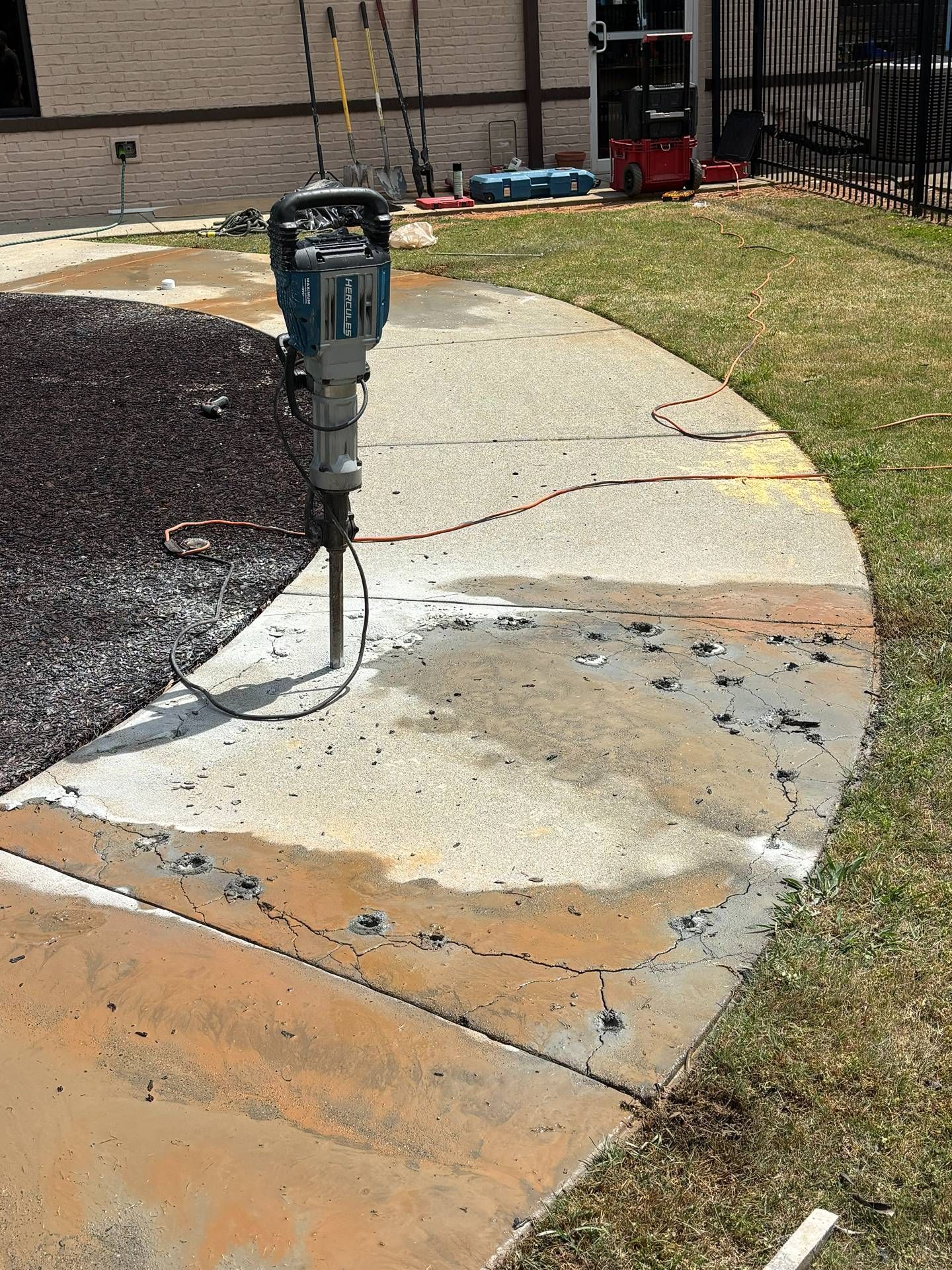 A jackhammer stands on a curved concrete walkway under repair, with fresh patches and dirt nearby.