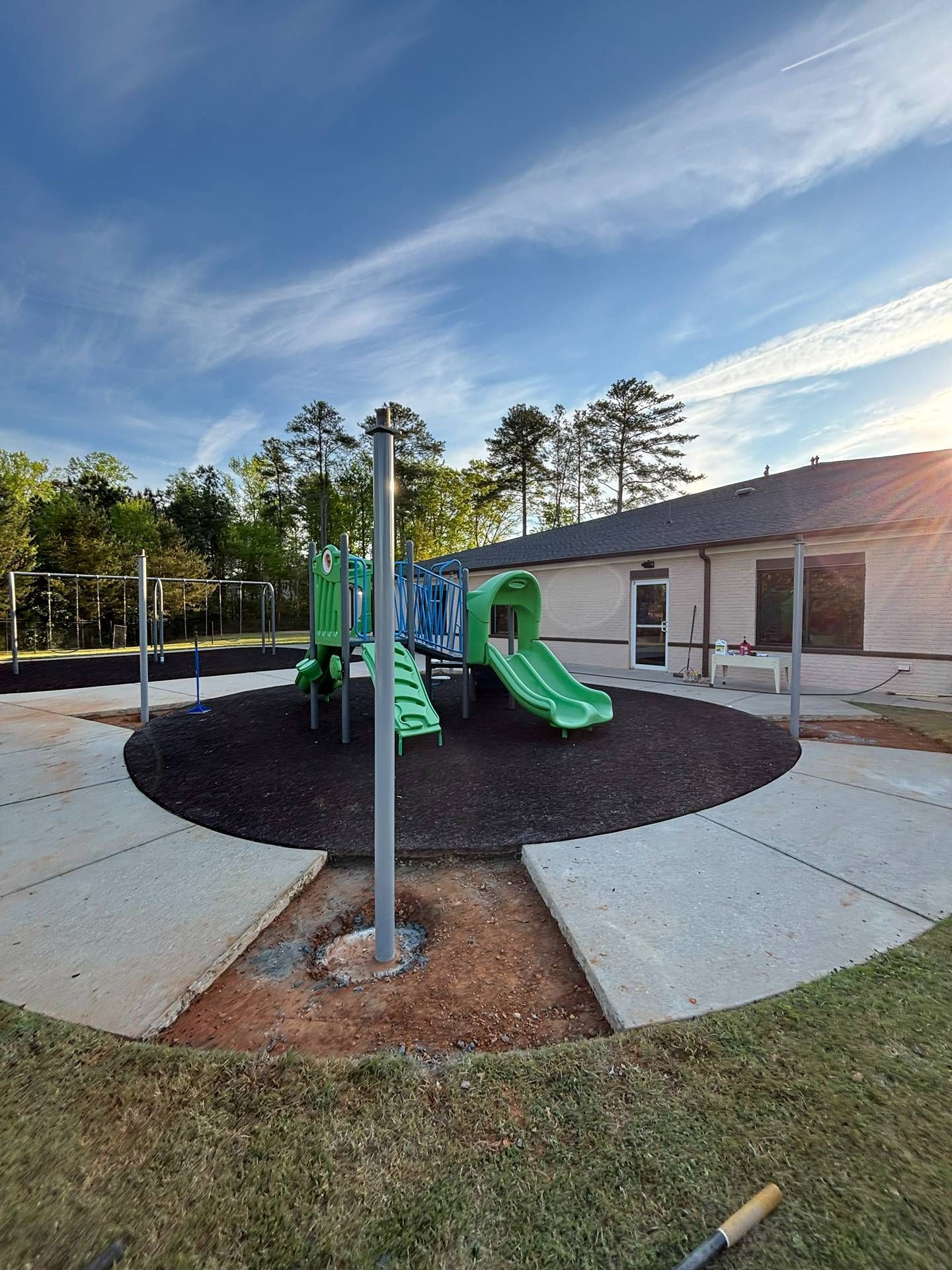 Small playground with green slides on a black rubber surface beside a building under a blue sky.