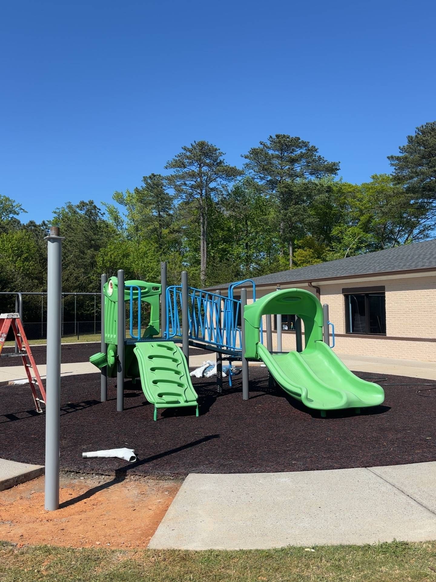 Green playground slide and climbing structure on mulch beside a brick building under a clear blue sky.