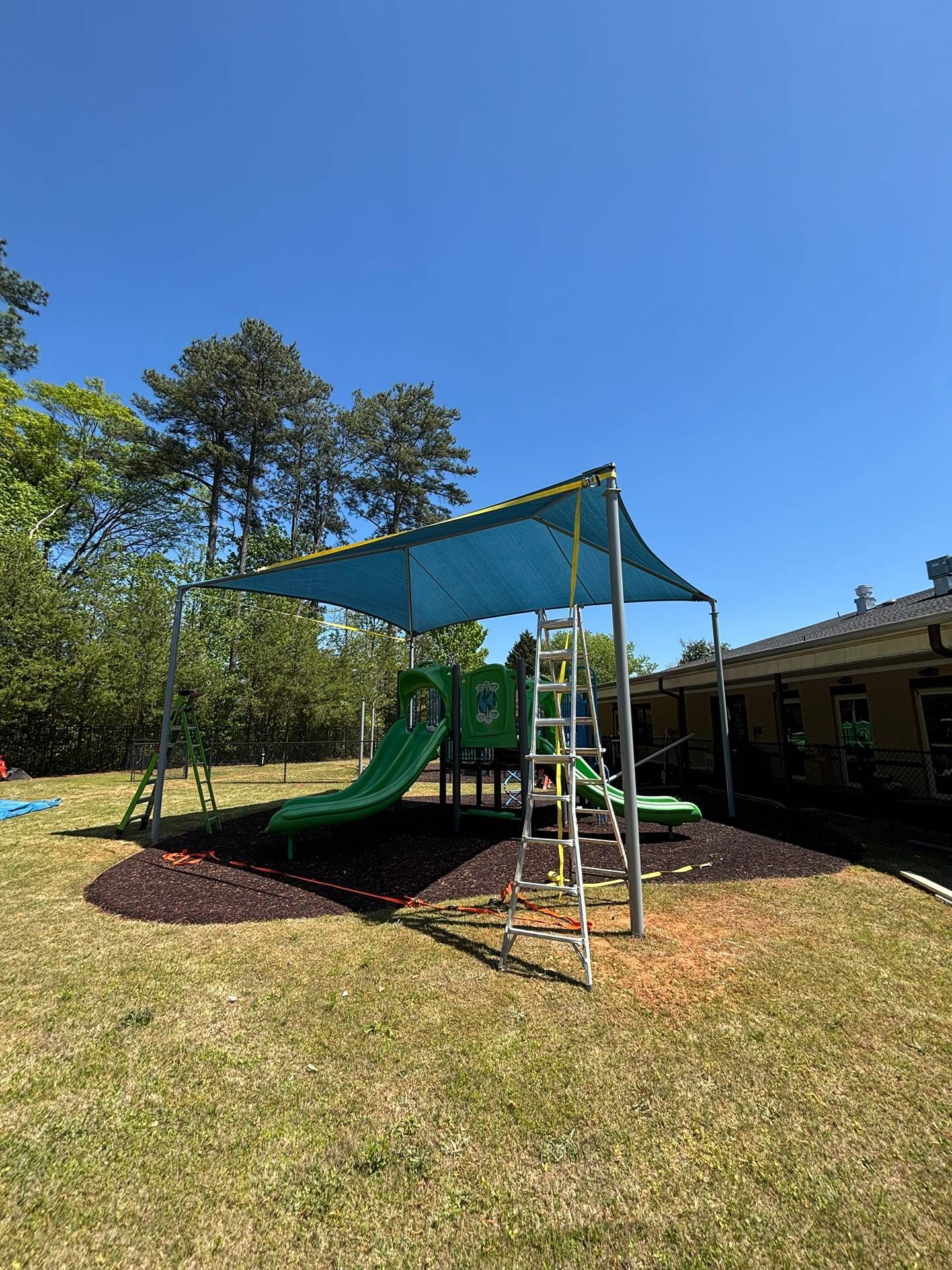 Outdoor playground with a green slide, ladder, and shade canopy on a grassy lawn under a blue sky