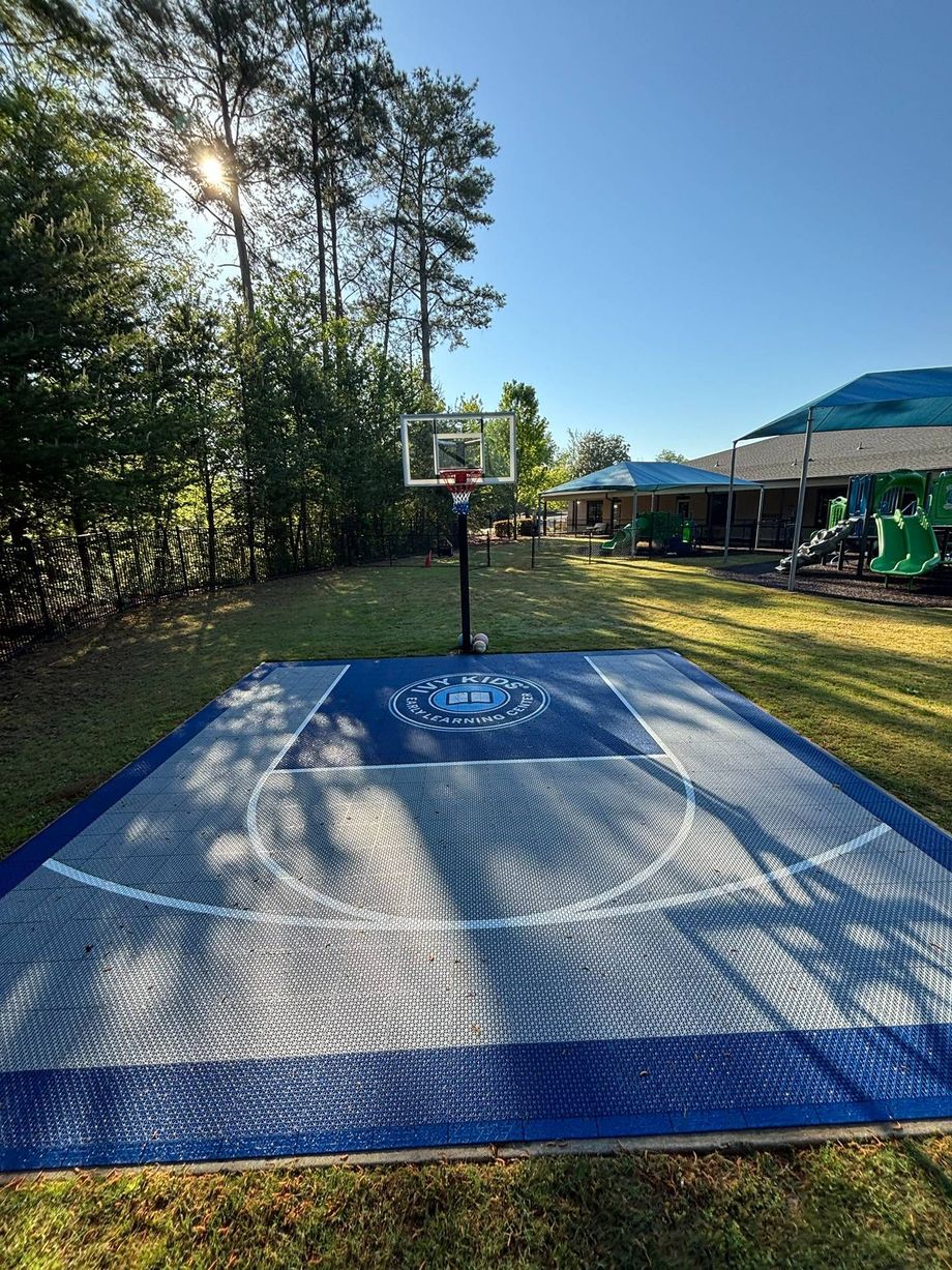 Backyard basketball hoop on a blue outdoor court, with trees and a house in the background.