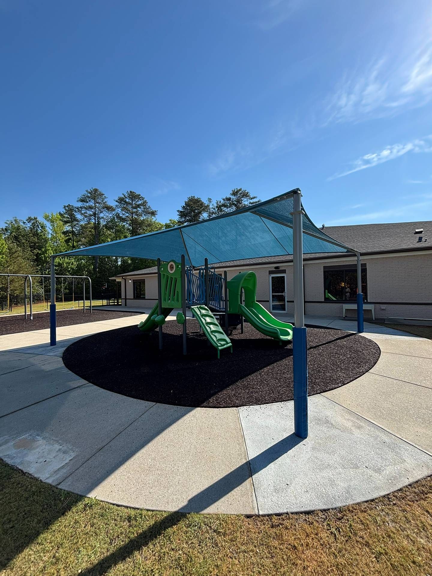 Covered playground structure with green slides and blue shade sails on a schoolyard court