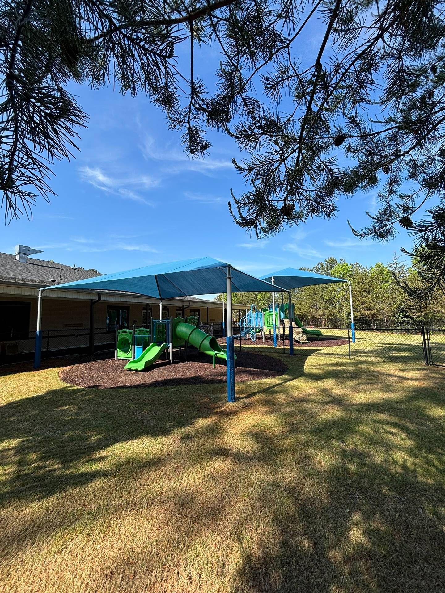 Shaded playground area with blue canopies, green play equipment, and a building in the background