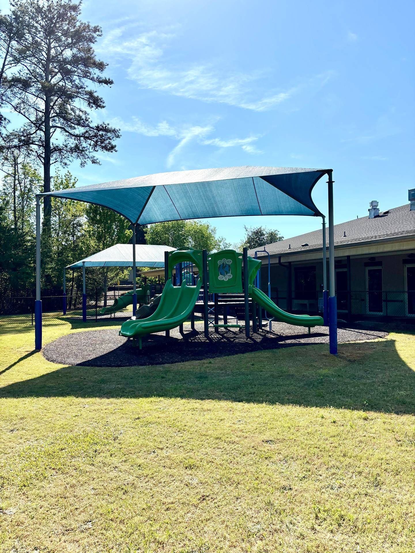 Playground with green slides under a shaded canopy beside a brick building on a sunny day