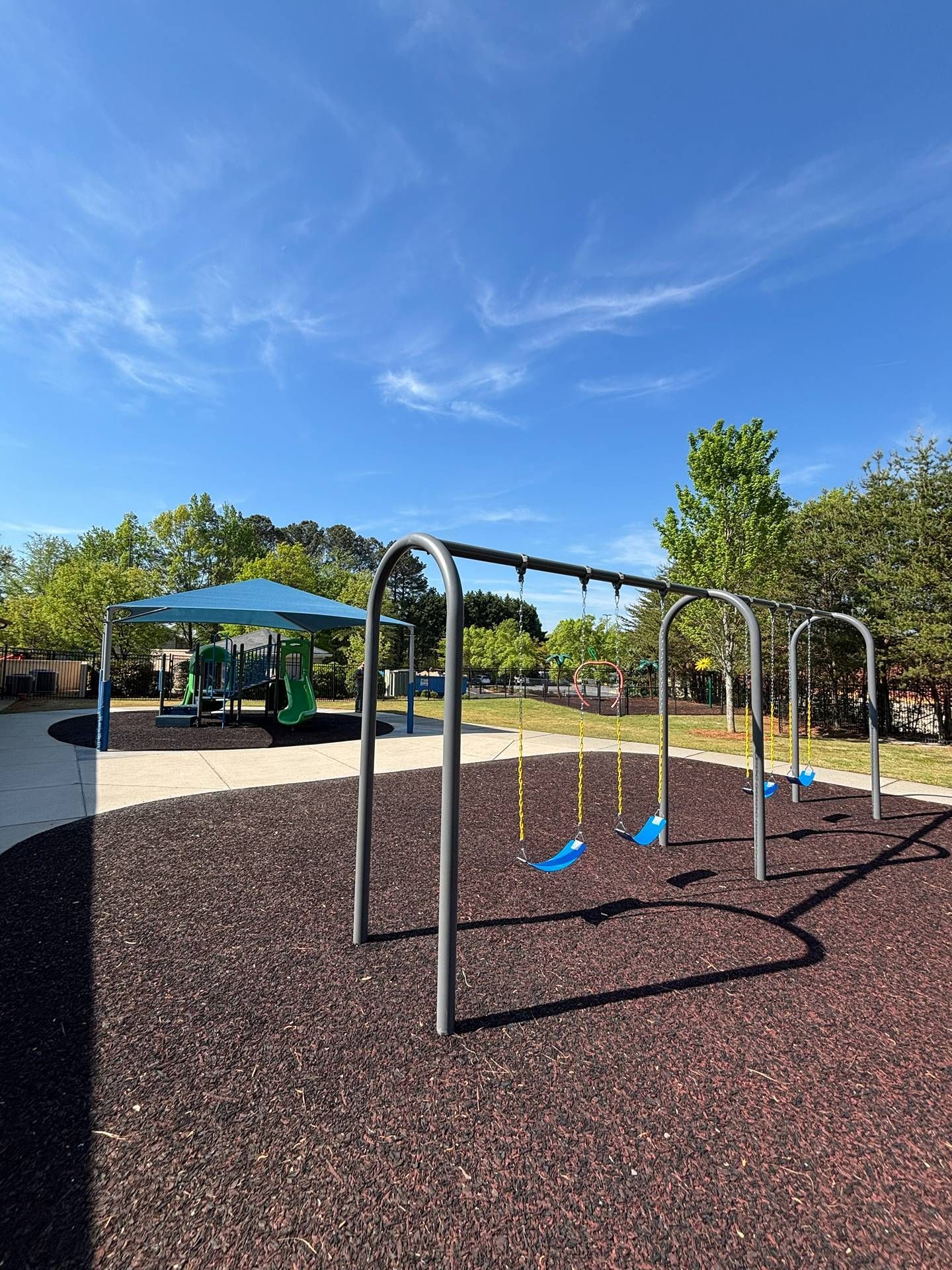 Playground with blue swings, metal climbing arches, and a shaded picnic area under a bright blue sky.