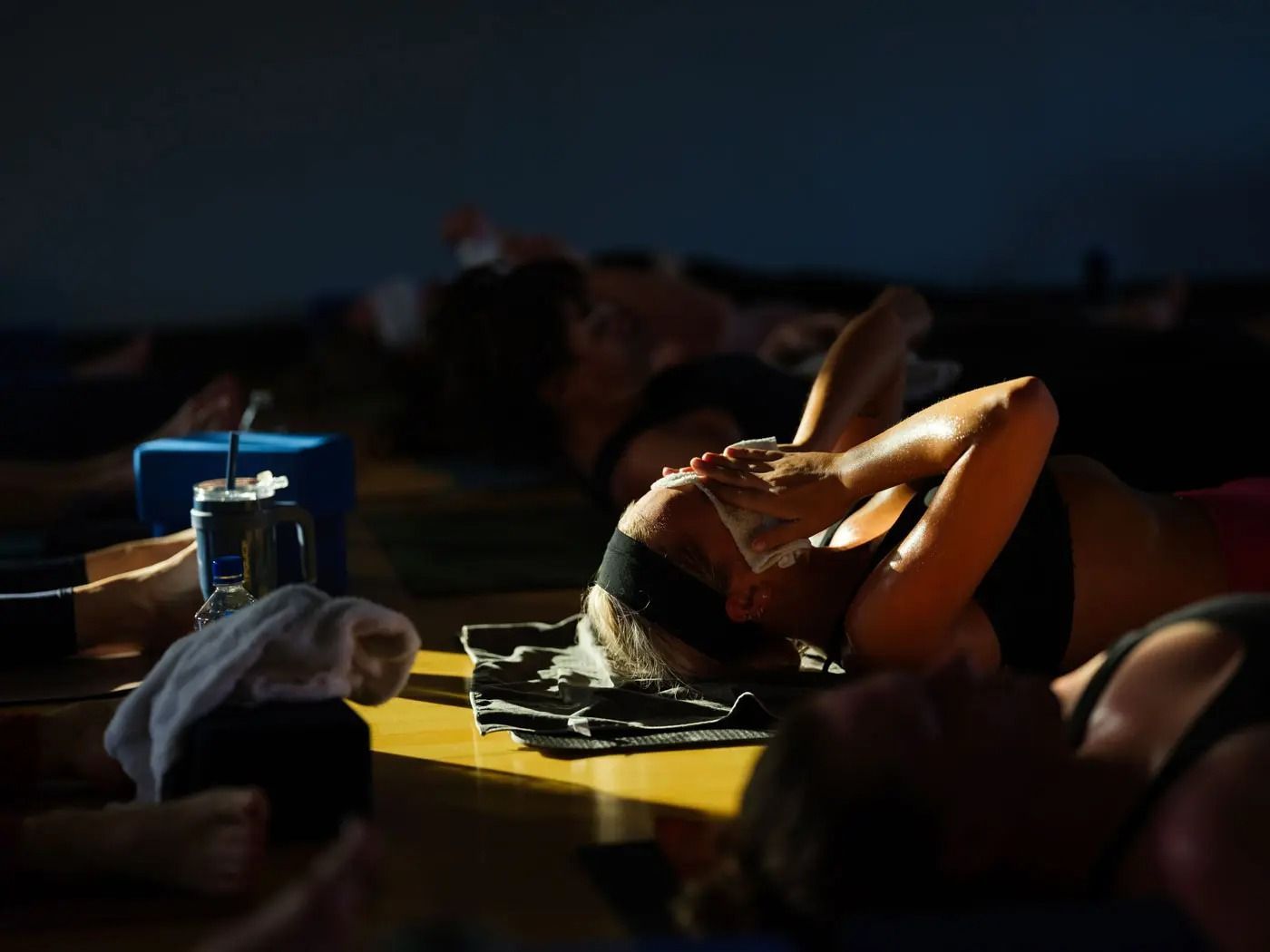 People lying on mats in a dimly lit room, with one person holding a cloth to their face in a bright beam of light.