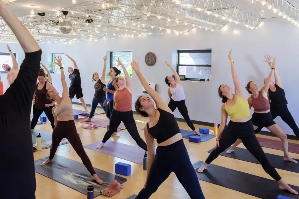 A group of people in a bright studio perform yoga, stretching into a side angle pose on individual mats.