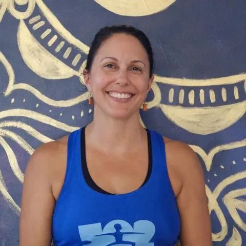 A smiling person wears a blue tank top, standing in front of a dark blue wall featuring gold, painted patterns.