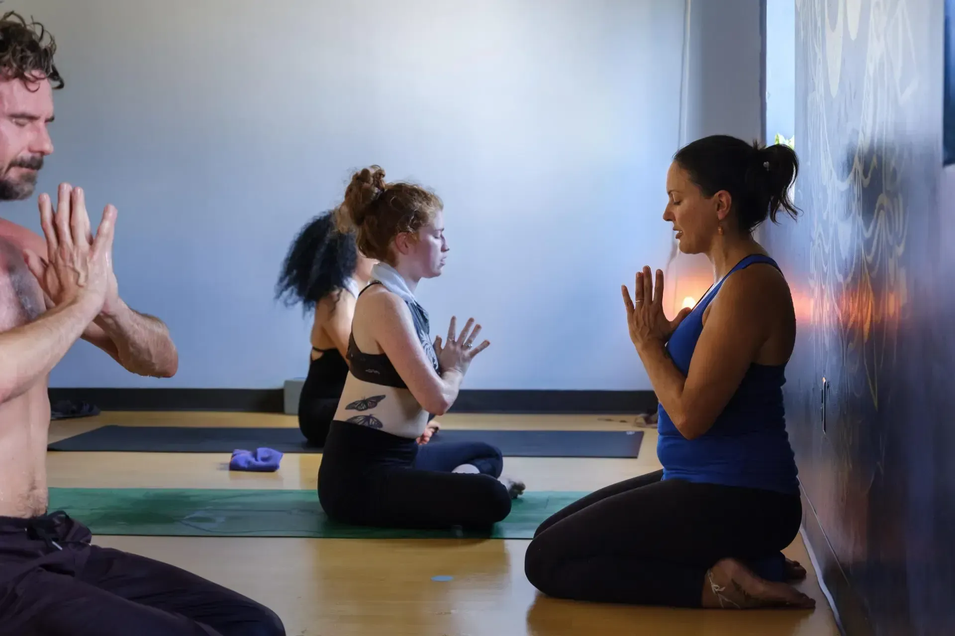 People in a studio sitting on mats with hands in prayer position, practicing yoga.