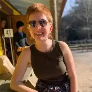 A person with short red hair and sunglasses smiles while sitting outdoors near a table with a number 41 sign.