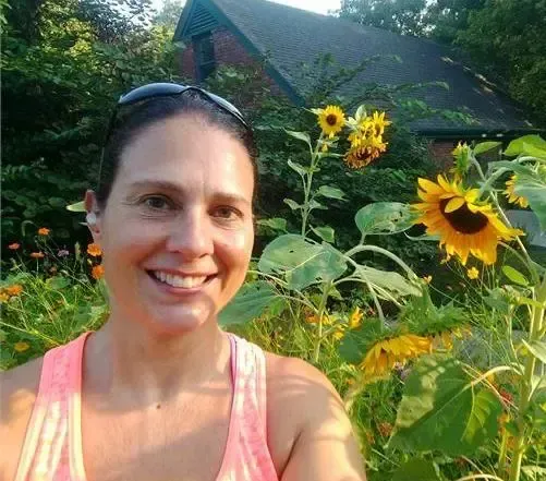 A woman wearing a pink tank top smiles in front of a garden with several yellow sunflowers and a brick house behind her.