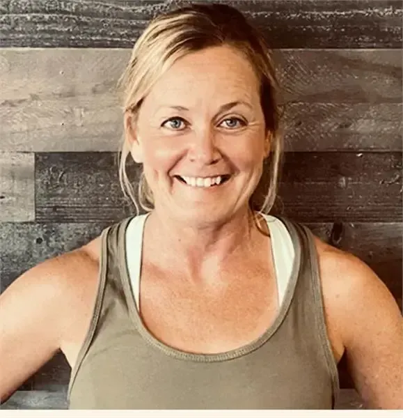 Smiling woman with blonde hair in a ponytail, wearing an olive green tank top against a dark horizontal wood wall.