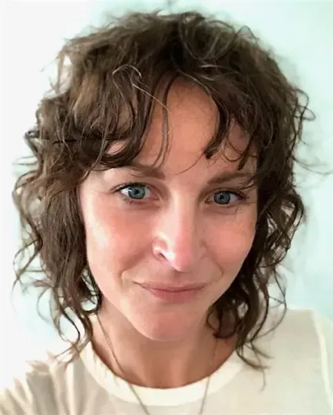 A person with curly brown hair and blue eyes looks at the camera with a gentle smile, wearing a cream-colored top.
