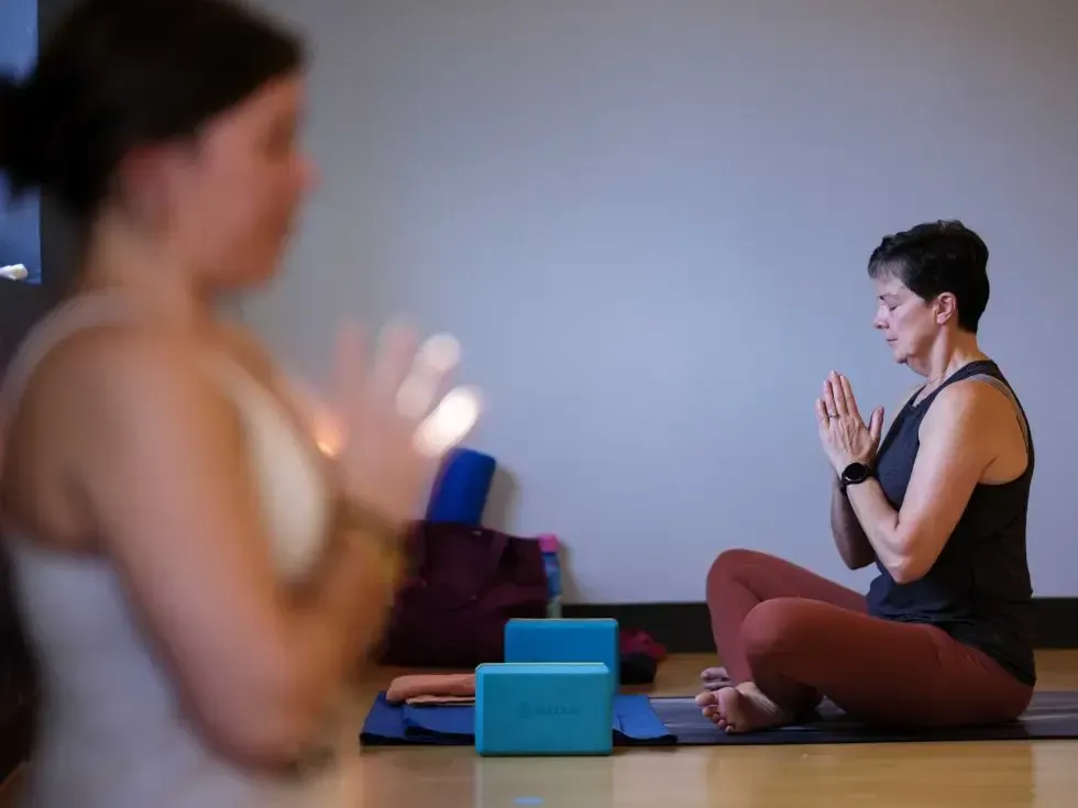 Two people sit in a yoga studio with hands in prayer position, focusing on meditation with yoga blocks nearby.