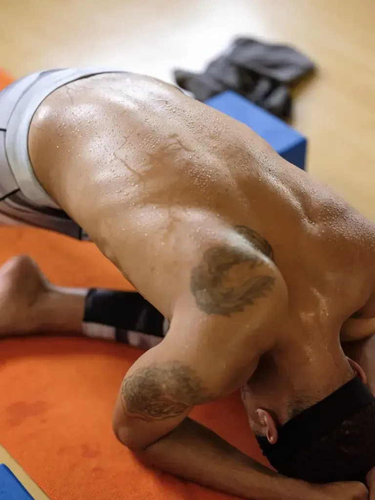 A sweaty person in athletic gear performs a child's pose on an orange mat in a gym.