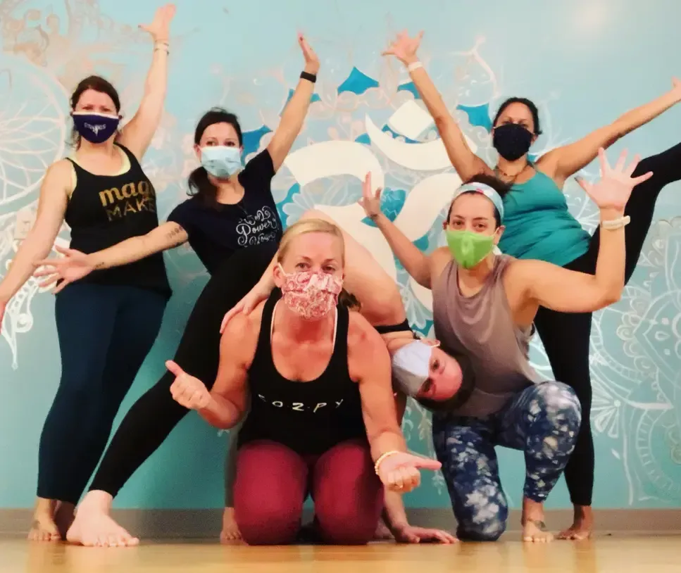 A group of six people in face masks pose playfully in a yoga studio with a mural of an Om symbol in the background.