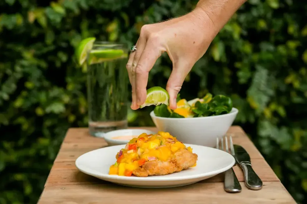 Hands hold a plate of seared salmon, yellow polenta cakes, roasted cherry tomatoes, mushrooms, and greens on purée.