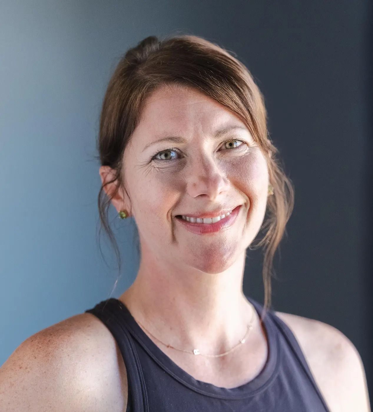 A person with shoulder-length hair tied back and a light dusting of freckles smiles gently against a blue background.