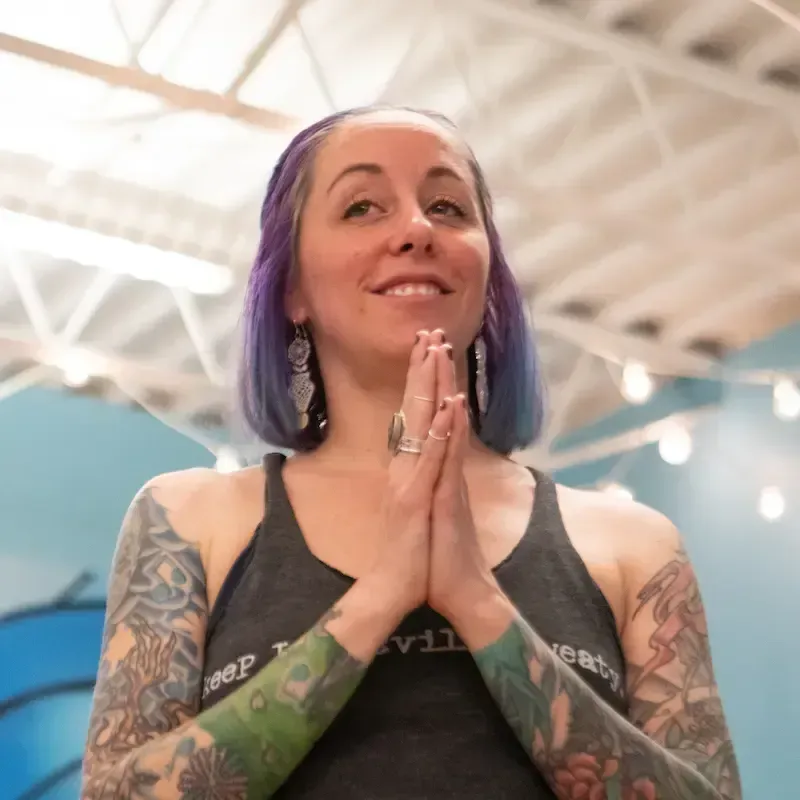 Smiling person with purple-blue hair and tattooed arms, hands clasped together indoors under bright lights in a yoga studio