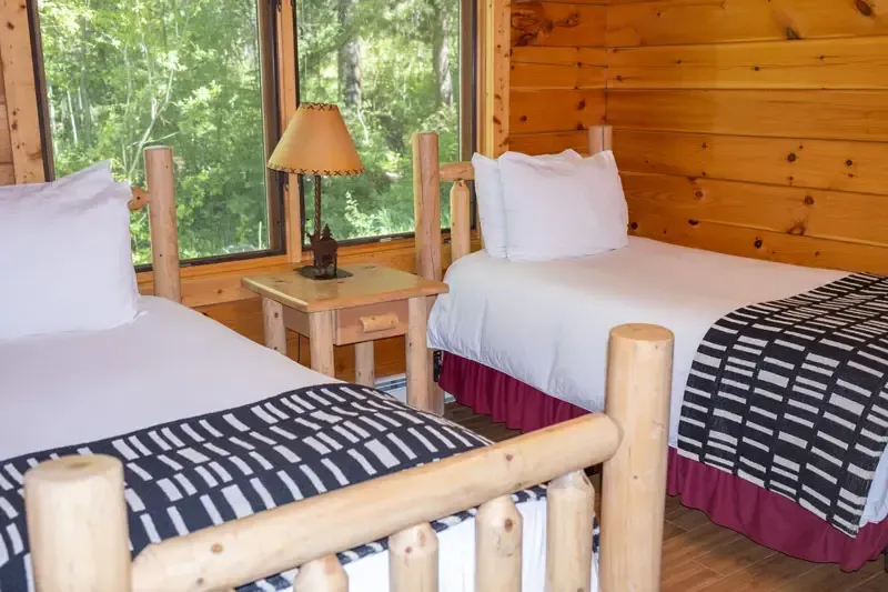 Two twin beds with white linens and patterned throws in a room with wood-paneled walls and a window.