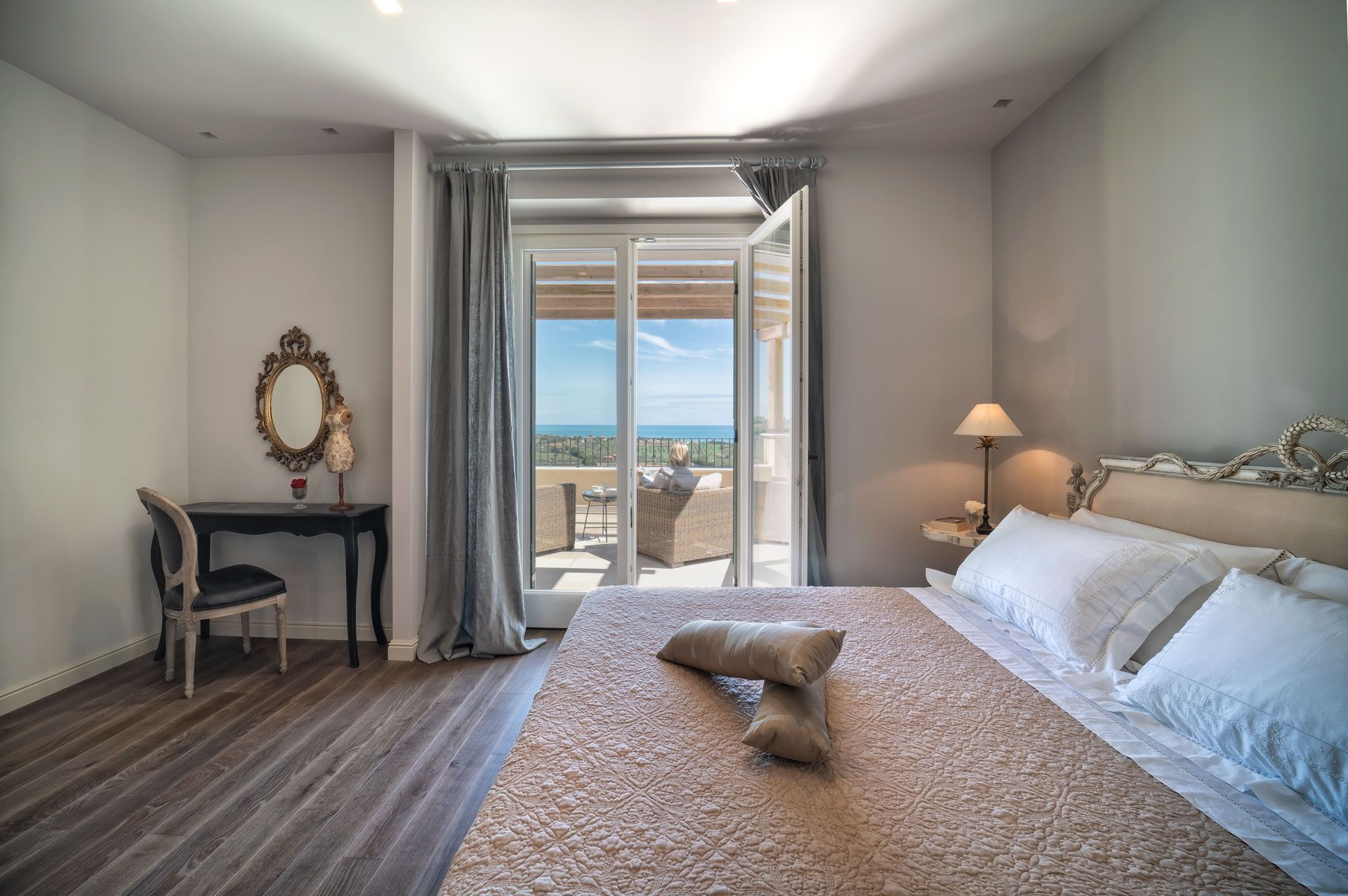 Bright bedroom with a bed, vanity, and open doors to a balcony overlooking the sea