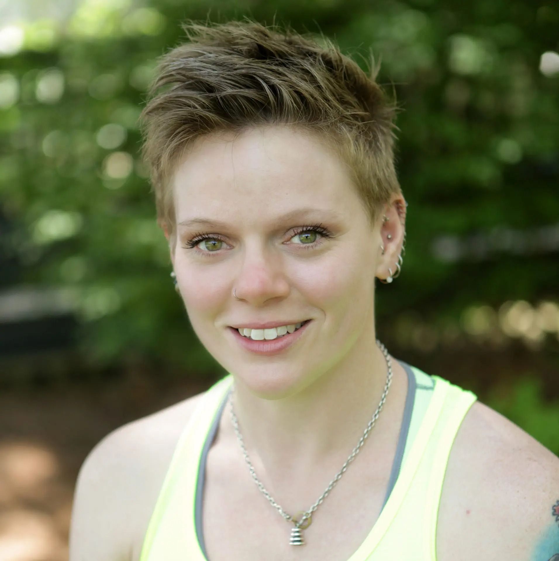 A person with a short, spiky hairstyle and a yellow tank top smiles in front of a green, out-of-focus background.