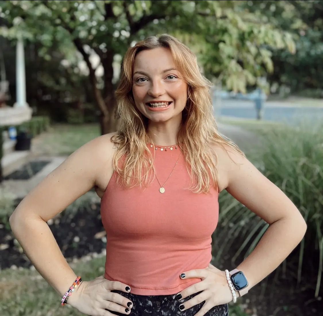 A smiling person with wavy blonde hair wears a coral tank top and necklaces, posing outdoors with hands on their hips.