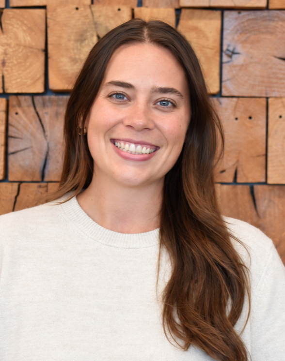 A person with long brown hair smiling against a plain, light-gray background while wearing a dark-colored shirt.