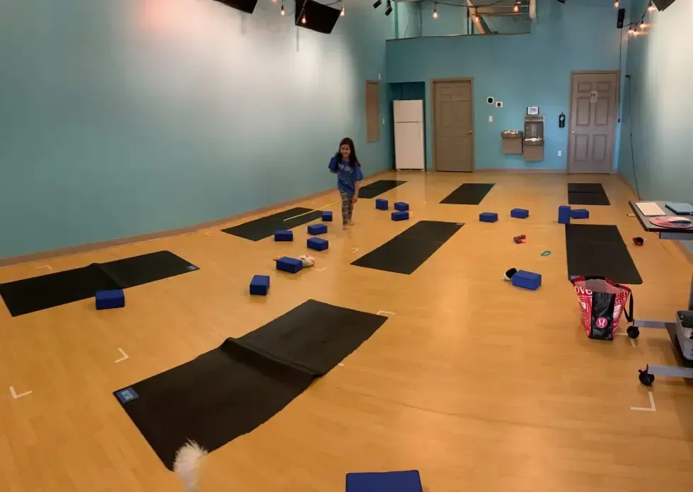 A person stands in a studio with teal walls and light wood floors, where black yoga mats and blue blocks are spread out.