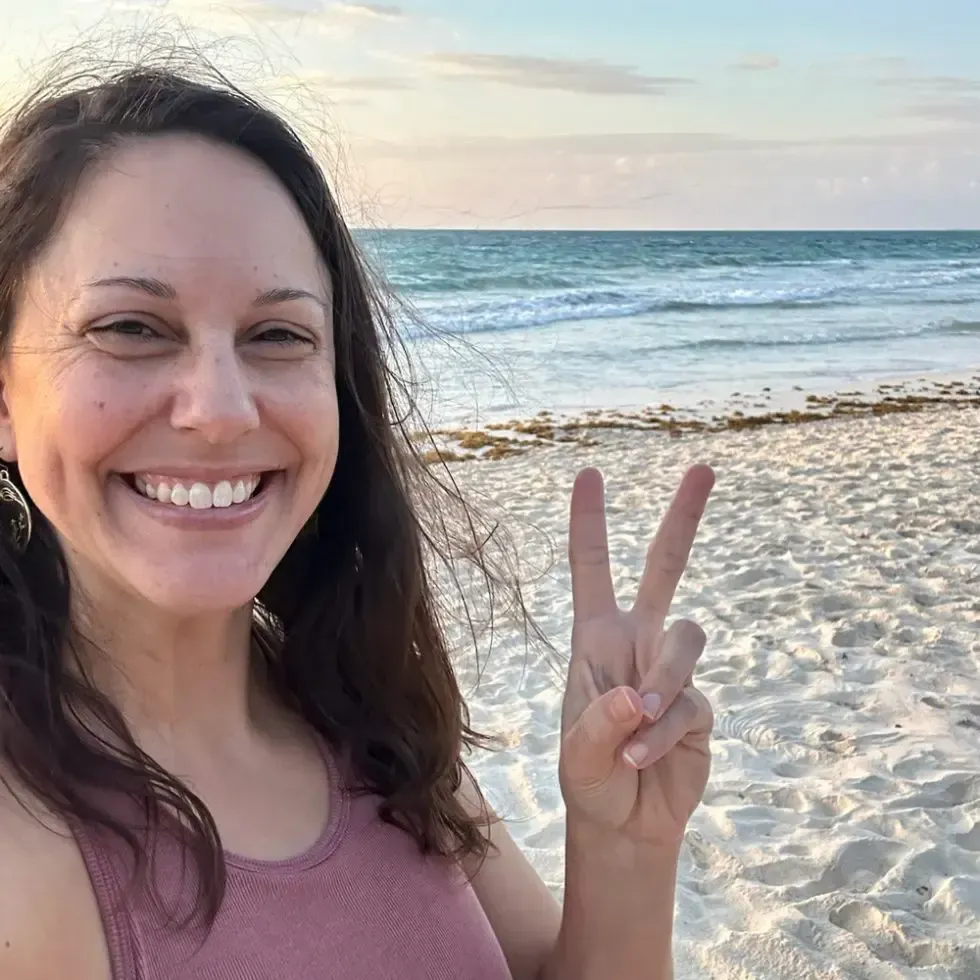 A smiling person in a pink top makes a peace sign with their hand on a sunny, sandy beach by the ocean.