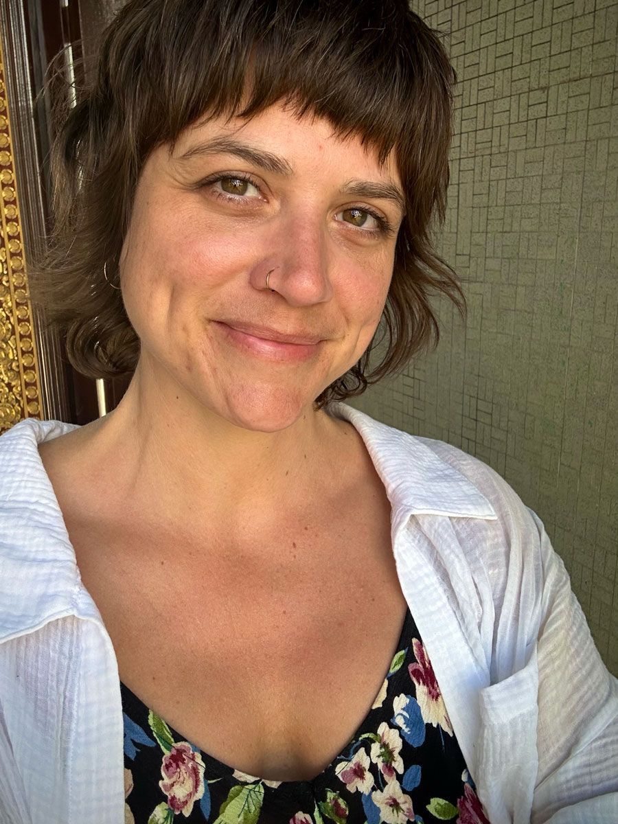 A smiling person with dark bangs and a nose ring, wearing a red patterned top against a textured pink background.
